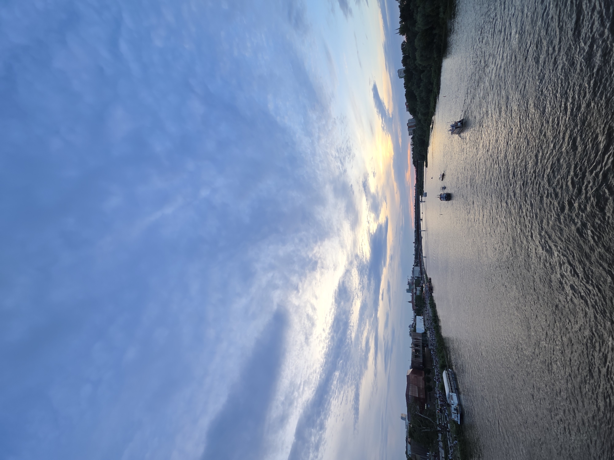The image captures a wide shot of the Vistula River in Warsaw, Poland, at dusk. The sky is a gradient of blues and grays with scattered clouds, transitioning to a soft yellow and orange glow near the horizon, indicating the setting sun. On the left bank, there is a line of buildings, including a modern structure with a reddish-brown facade, and a long white boat docked along the embankment. A crowd of people is visible on the riverside promenade. Several small boats are navigating the river, one of which is in the foreground on the right, moving away from the viewer. The right bank is densely covered with trees, with some buildings visible peeking through. The river's surface is rippled, reflecting the subdued light of the sky.