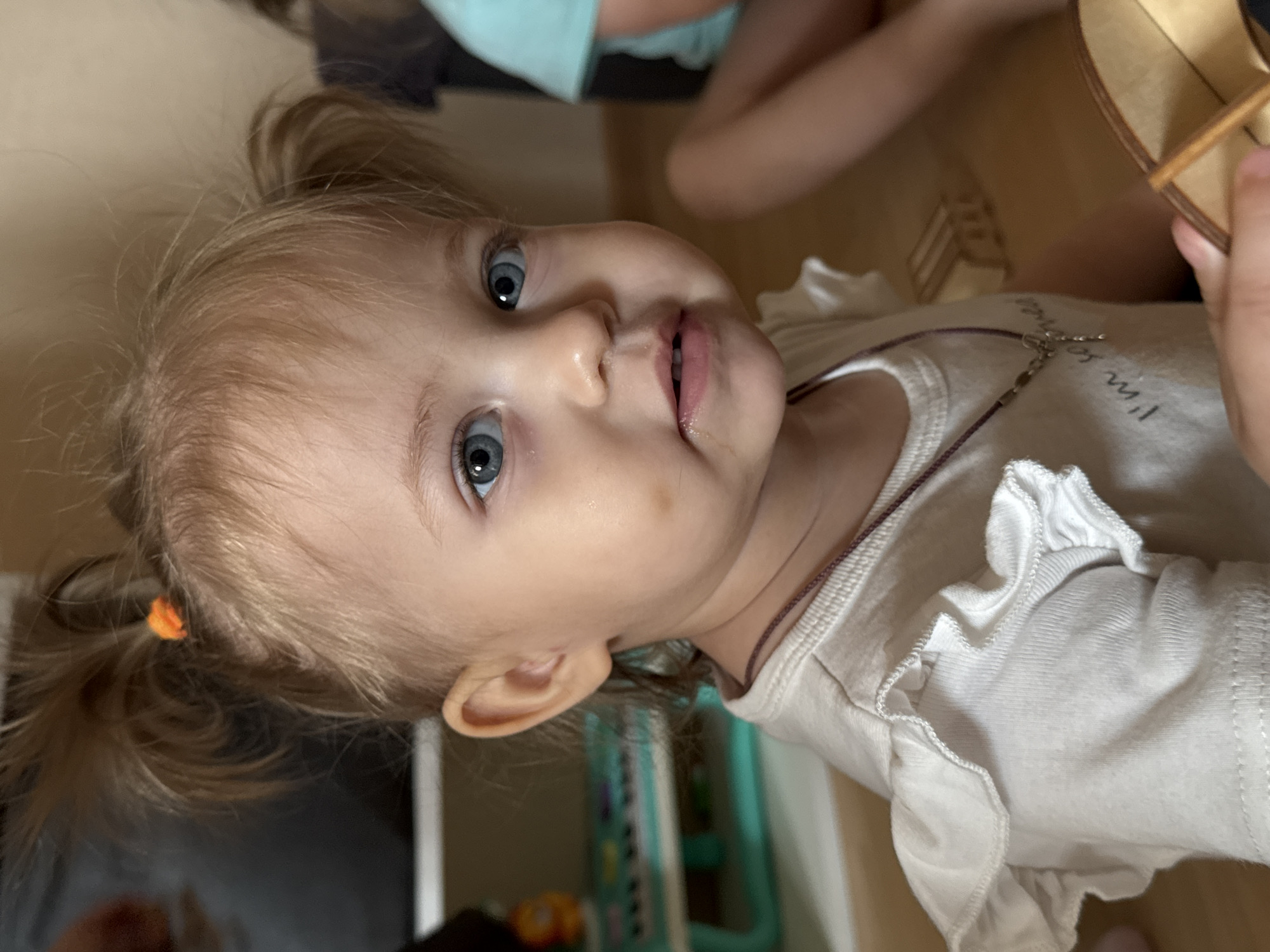 The image shows a close-up, eye-level shot of a young Caucasian girl with blonde hair in pigtails, wearing a white ruffled shirt and a necklace. She has blue eyes and a small dark spot on her left cheek. The background is blurred and shows a room with wooden floors and a shelf with toys. The girl is looking directly at the camera with her mouth slightly open. No specific location in Chaikovsky, Russia, can be identified from the image.
