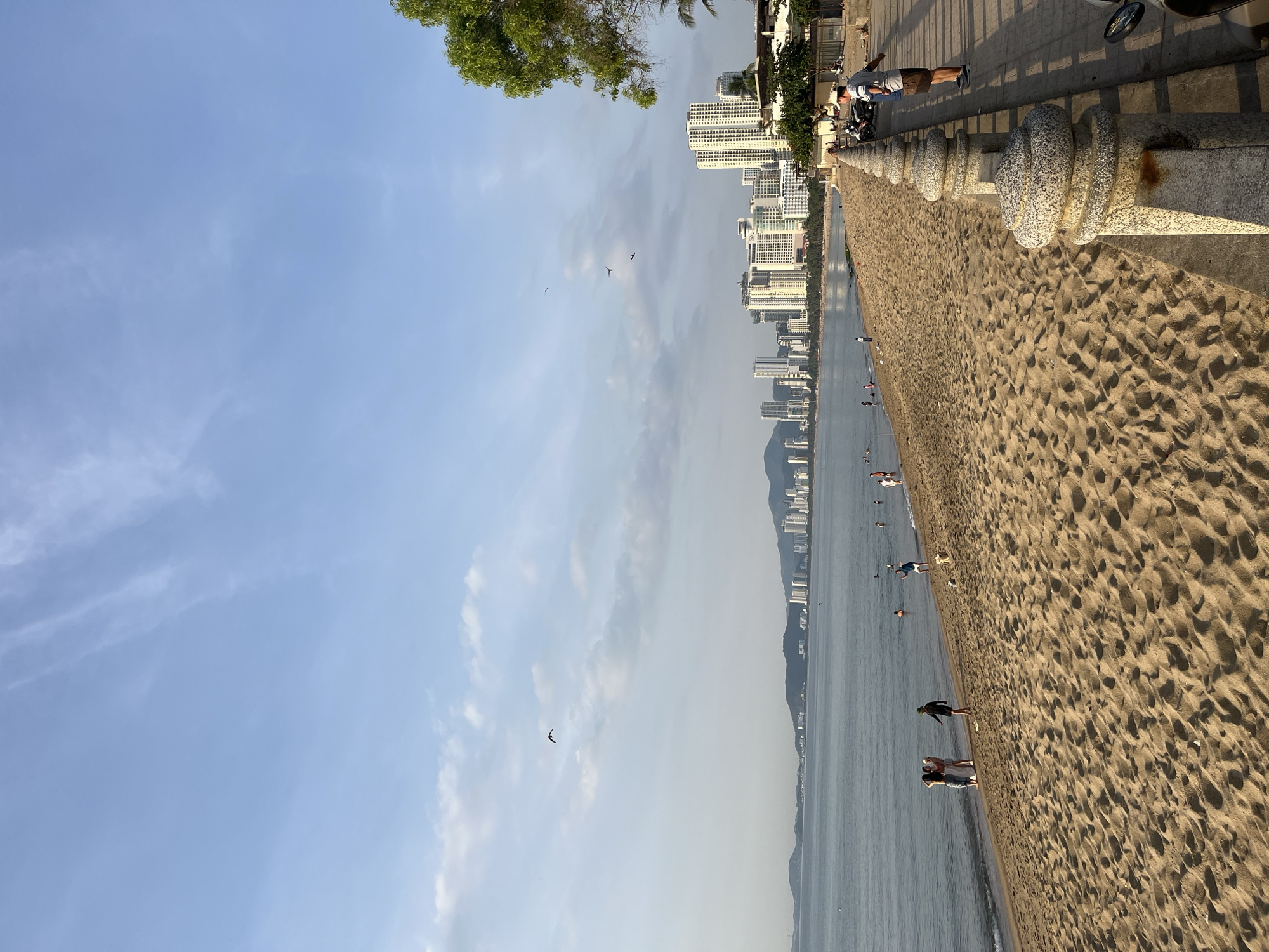 The image depicts a beach in Nha Trang Ward, Vietnam. A sandy beach stretches along the coastline, bordered by a concrete walkway with a decorative railing. In the distance, a city skyline with tall buildings can be seen. People are visible on the beach and in the water. The sky is blue with some clouds.