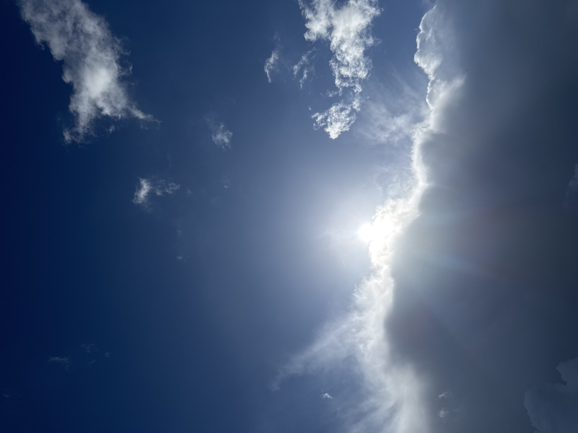 A bright sun is obscured by a large, dark cloud, casting rays of light downwards in a celestial display over a city. The sky is a deep blue, with wispy white clouds scattered across its expanse. The dark cloud dominates the lower half of the image, its edges illuminated by the hidden sun.