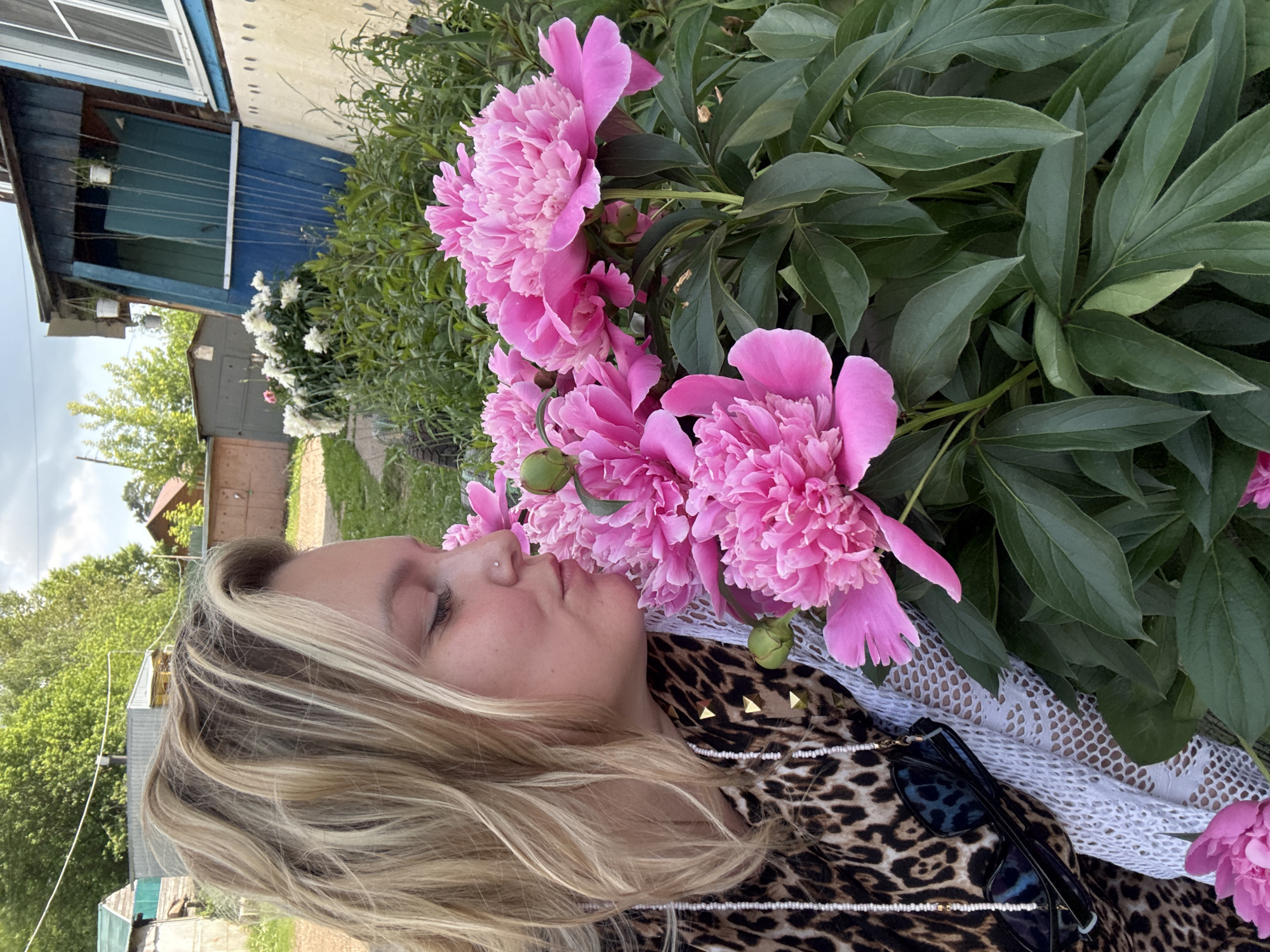 A woman with blonde hair is smelling pink peonies in a yard in Chaikovsky, Russia. She is wearing a leopard print top and a white lace scarf. Sunglasses hang from a beaded strap around her neck. In the background, there is a wooden house with blue shutters and other buildings. The area is surrounded by trees and greenery.