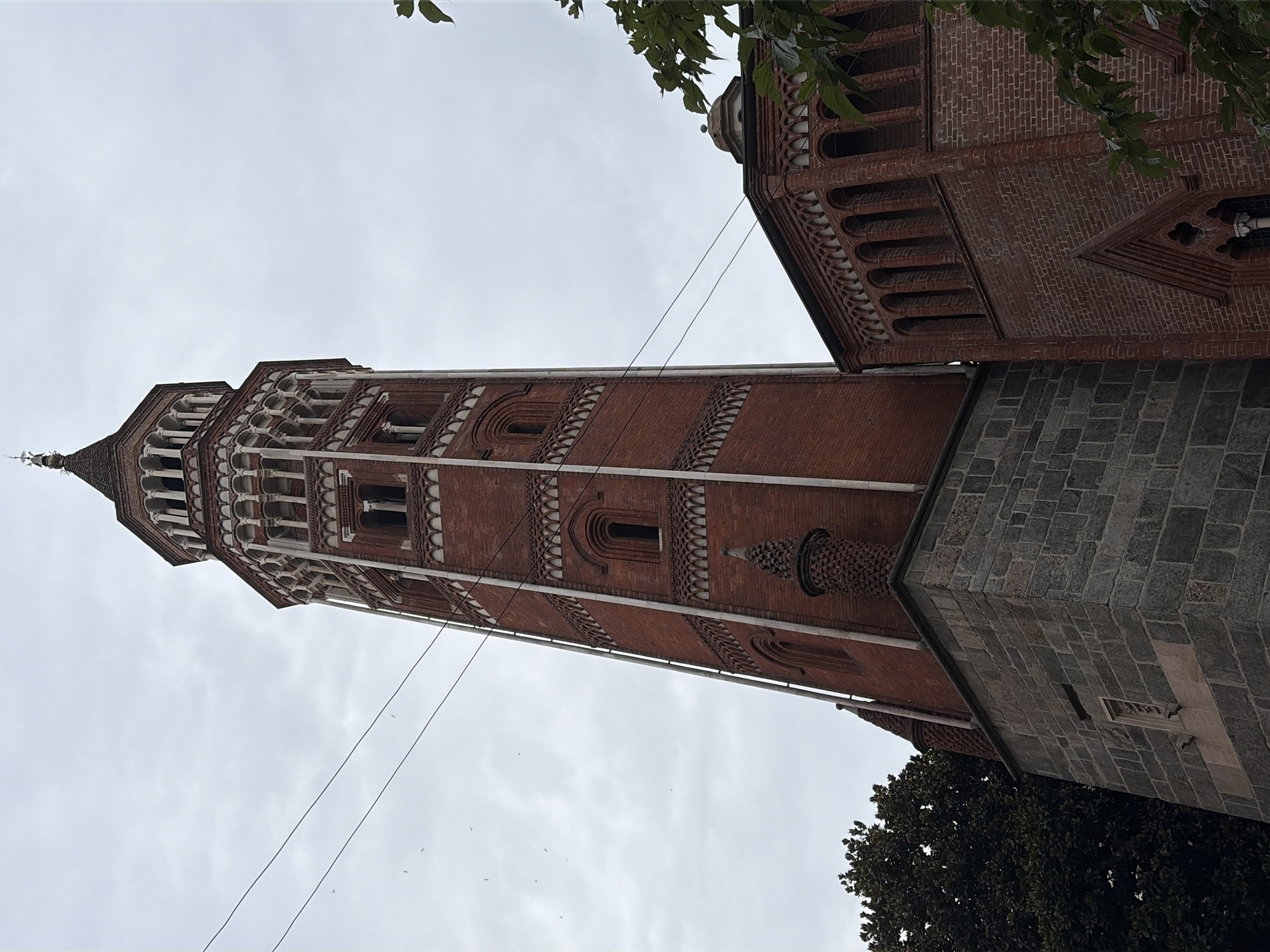 The image shows a view of the Basilica di Sant'Ambrogio in Milan, Italy, focusing on its iconic bell tower, the Campanile dei Canonici. The tower is constructed primarily of red brick with white stone accents, featuring multiple levels of arched windows and decorative cornices. The lower section of the tower is made of grey stone blocks. Adjacent to the tower is another brick structure, part of the basilica, with similar arched detailing. The sky is overcast and cloudy. Several thin wires are visible crossing the sky in front of the tower. A dark green tree is partially visible on the left, and green leaves frame the right side of the image. Small birds are scattered in the sky.