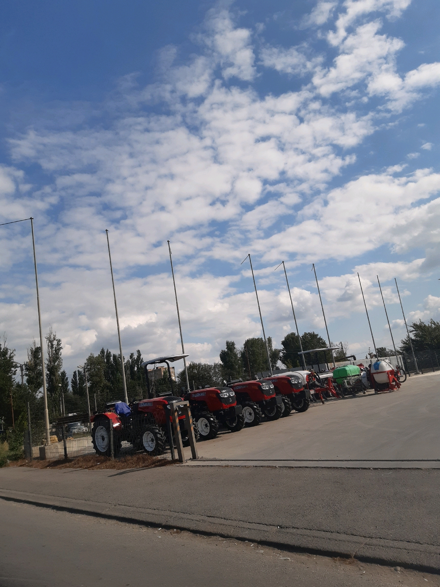 A low-angle, eye-level, wide shot captures a clear blue sky with scattered white clouds above a row of tractors and agricultural equipment parked in an outdoor lot. The lot appears to be paved with asphalt. Several tall, thin poles with flags at the top are spaced evenly across the scene, extending from the foreground to the background. On the left, a metal fence separates the lot from an area with trees and vegetation. The tractors are red and black with large wheels. One tractor in the foreground has a blue tarp draped over its seat. Behind the tractors, there is green and white agricultural machinery. In the distance, the tops of buildings and more trees are visible. The overall scene is of a lot displaying agricultural machinery under a partly cloudy sky.