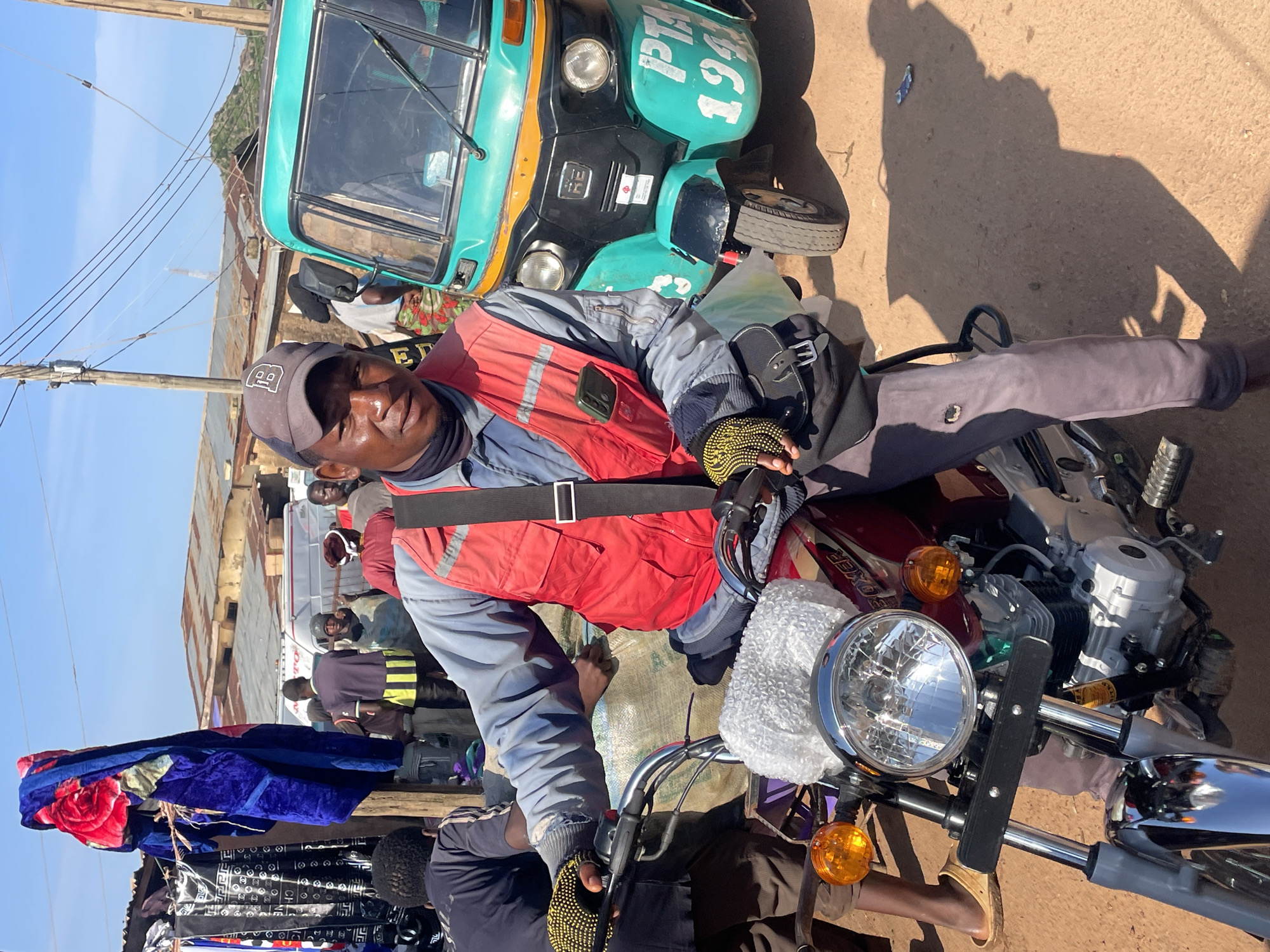 In Gura Top, Nigeria, a man wearing a red vest, dark blue shirt, and grey trousers is seated on a red motorcycle. He has on black gloves with yellow accents and a black baseball cap. Next to him, a green and yellow auto rickshaw is parked. Behind them, a market scene unfolds with stalls displaying clothing and blankets, tin-roofed buildings, and a clear sky. The ground is dirt.