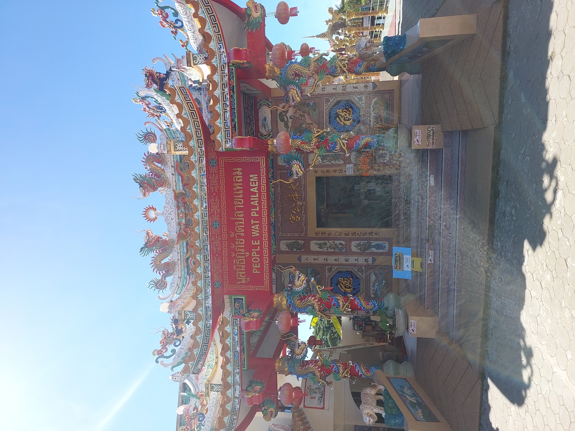 The image shows the entrance to the People Wat Plai Laem temple, a prominent landmark in Baan Plai Laem, Thailand. The temple features a vibrant, ornate architectural style, characteristic of Chinese temples, with intricate dragon motifs adorning the roof and pillars. The main entrance is flanked by two large, colorful dragon sculptures wrapped around red pillars. Above the entrance, a red banner displays text in both Thai and English, identifying the temple as "PEOPLE WAT PLAILAEM". Red lanterns are hung, adding to the festive atmosphere.
In front of the temple, a wide set of stairs leads up to the entrance. To the left of the stairs, a white tiger statue is visible, positioned on a raised platform decorated with lotus flowers. To the right of the stairs, another tiger statue is partially visible. Various signs, some with Thai text and iconography, are placed near the stairs, including one that appears to be a prohibition sign with a shoe symbol, suggesting a requirement to remove footwear before entering. The ground in front of the temple is paved with hexagonal stones, and shadows from the structure and sunlight create a dappled effect. The sky above is clear and bright blue. The overall scene conveys a sense of cultural and religious significance, with a strong emphasis on traditional Chinese artistic elements.