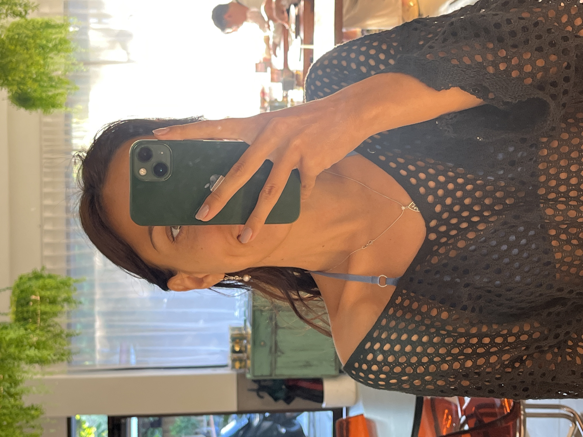 A woman is captured taking a mirror selfie, holding a dark green smartphone (likely an iPhone) with her right hand. Her face is partially visible in the reflection, showing one eye looking towards the phone, with dark hair, and an ear adorned with a pearl earring. She is wearing a black mesh top, under which a blue strap, possibly from a bra or swimsuit, is visible. A delicate silver necklace with a small, possibly heart-shaped, charm hangs around her neck. Her nails are neatly manicured with a light, natural-colored polish.
The background reflection reveals a bright, sunlit indoor setting. Through a window, white horizontal blinds and a lush green plant are visible. A blurry figure, appearing to be another person, is also reflected further back, suggesting a public or commercial space like a salon or shop. The overall impression is a well-lit, daytime scene. The location context is Kuta Selatan, Indonesia. There is no discernible text visible in the image.