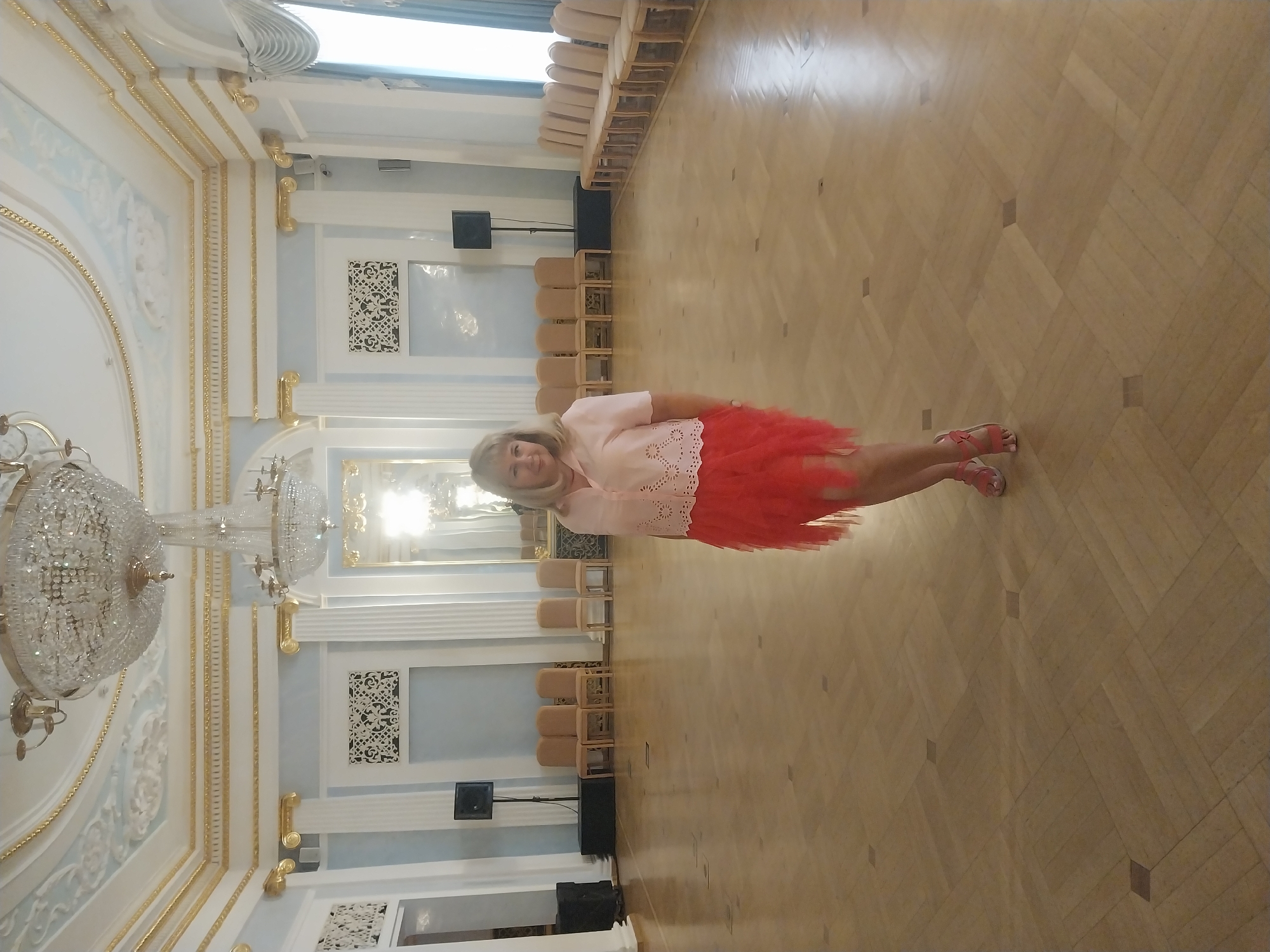 A woman stands in a grand ballroom, her light pink blouse and ruffled coral skirt creating a vibrant contrast against the pale blue walls and ornate gold trim. Chandeliers hang from the high ceiling, illuminating the polished wooden floor. Rows of chairs line the walls, and tall columns support the elaborately decorated architecture. She wears pink sandals.