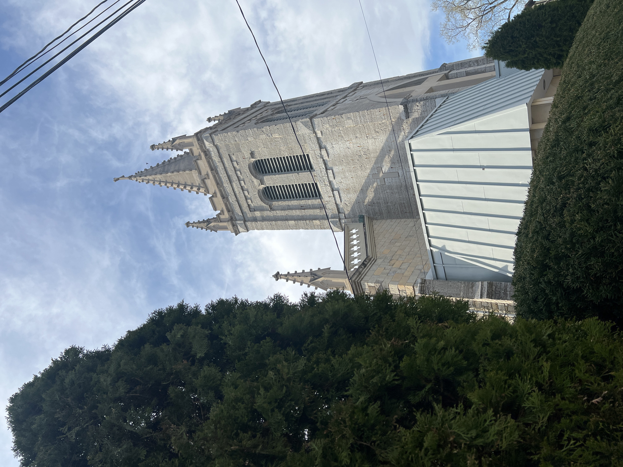 This image shows a view of the bell tower of St. George's Church in Newburgh, New York, United States. The tower is constructed of grey stone and features arched windows with louvers. Decorative spires adorn the top of the tower. A modern addition with a light grey, metal roof is attached to the side of the church. Evergreen trees partially obscure the base of the building. Power lines are visible against a cloudy sky.