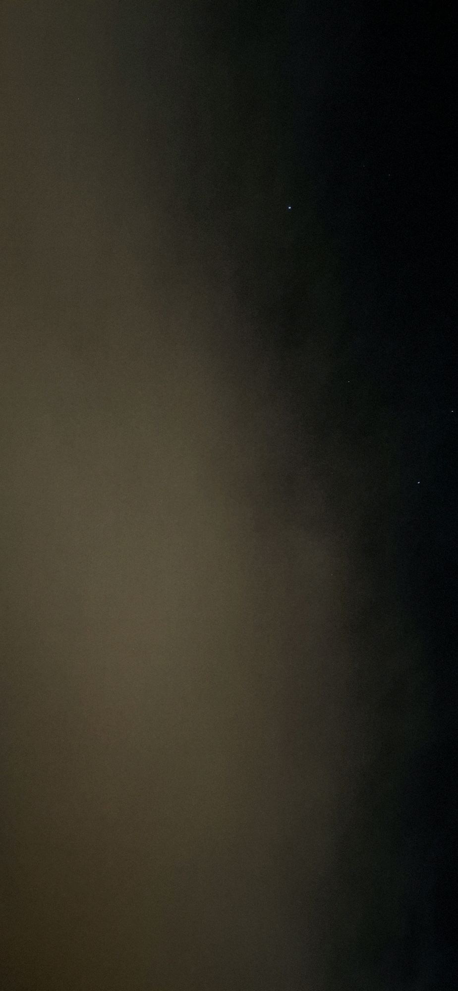 The image depicts a night sky filled with a scattering of stars, some of which are visible through a hazy atmosphere. The sky transitions from a lighter, brownish hue on the left to a darker, black hue on the right, suggesting either atmospheric conditions or a gradient of light pollution. One prominent star, appearing as a bright blueish dot, is situated in the upper right quadrant of the image. Another, fainter star is visible slightly to the left and below it. Further down and to the right, a cluster of three tiny stars is discernible. The image was captured in Yerocham, Israel, likely outdoors, with the focus on the celestial display. No people, animals, or man-made structures are visible in the frame.