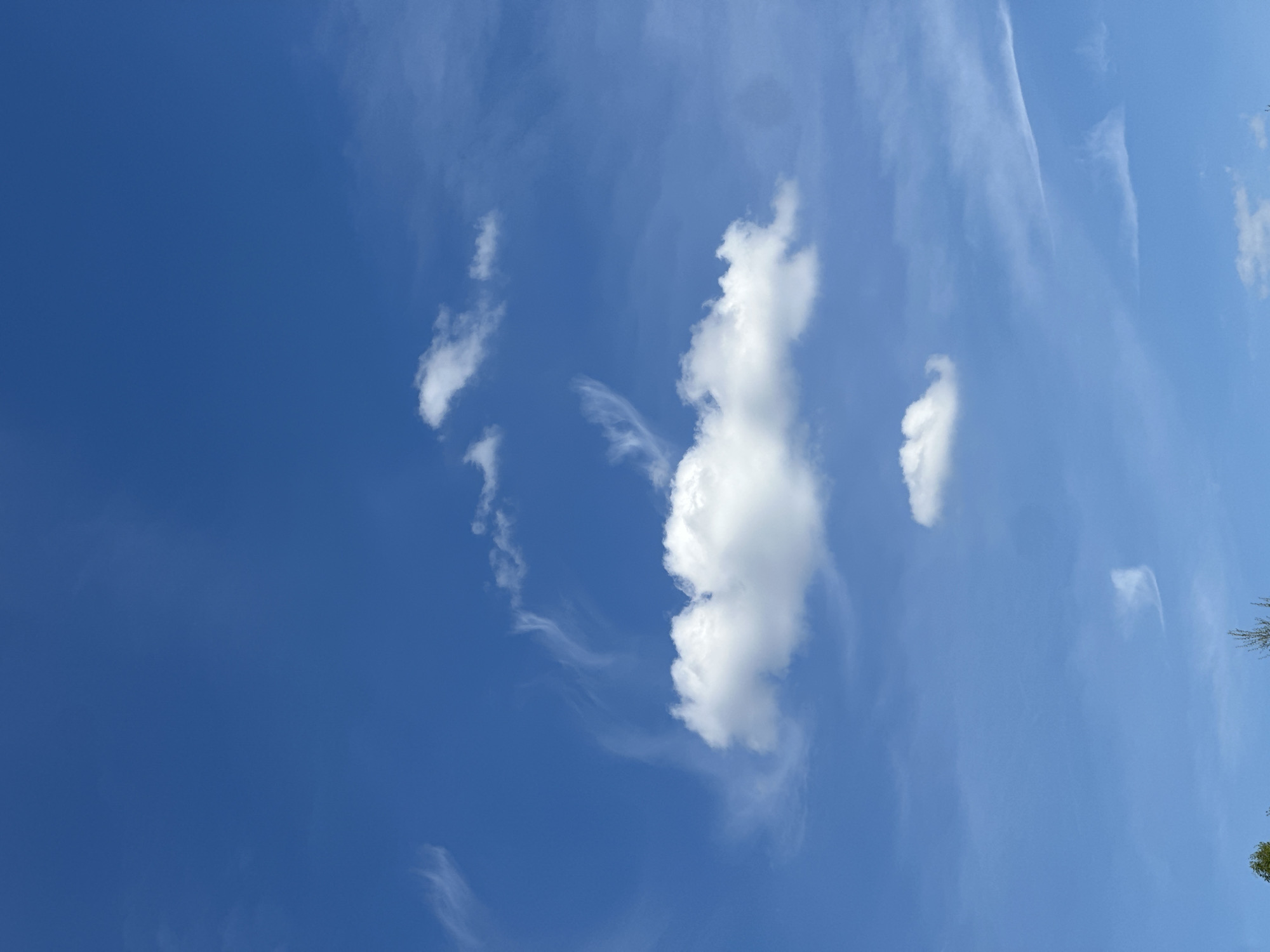 The image shows a clear blue sky with several clouds. Some of the clouds are wispy and thin, while others are larger and more cumulus-like. The clouds are scattered across the sky. The photo appears to be taken outdoors during the daytime. The location is Unknown, Unknown.