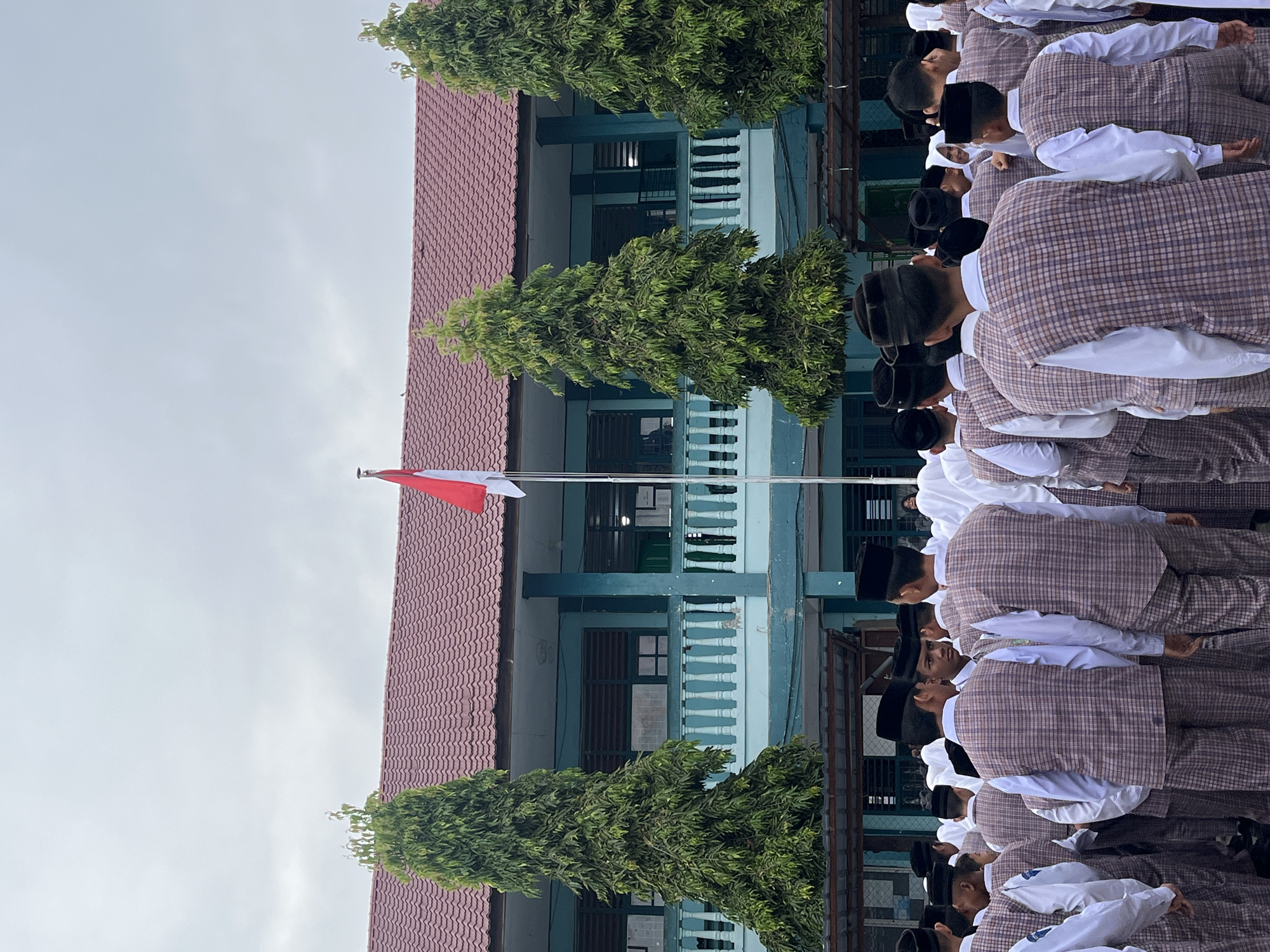 In Lhokseumawe, Indonesia, a group of students, primarily boys, stand in formation facing a school building. They are dressed in white shirts, plaid vests, and black caps, suggesting a school uniform or religious attire. Many are also wearing white headscarves. In front of the building, a flag pole with the Indonesian flag (red and white) is visible, indicating a flag-raising ceremony or assembly. The school building itself is light blue with a red tiled roof and a balcony with white railings. Tall, slender green trees flank the building, and the sky is overcast. The overall scene suggests an organized outdoor school event, likely a flag ceremony.