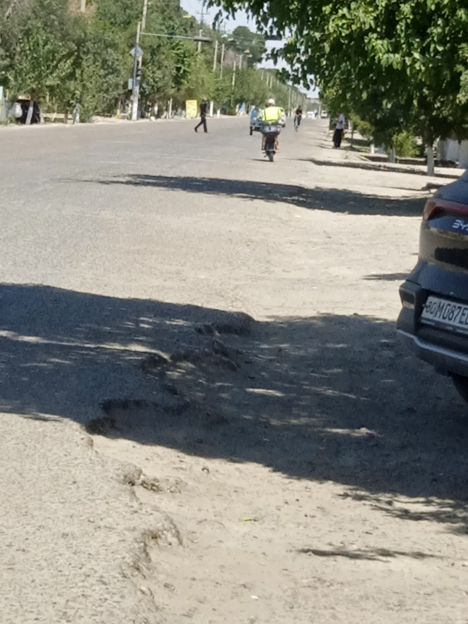 A medium shot shows a street scene with a road that has noticeable potholes. In the foreground, the edge of a dark-colored car, possibly an SUV, is visible on the right side, with its license plate reading "80 MG087ED". In the background, a person is riding a motorcycle, wearing a bright yellow vest. Several other pedestrians and vehicles are present further down the road, with trees lining the sides. The lighting suggests it is daytime, with shadows indicating the sun is casting light.