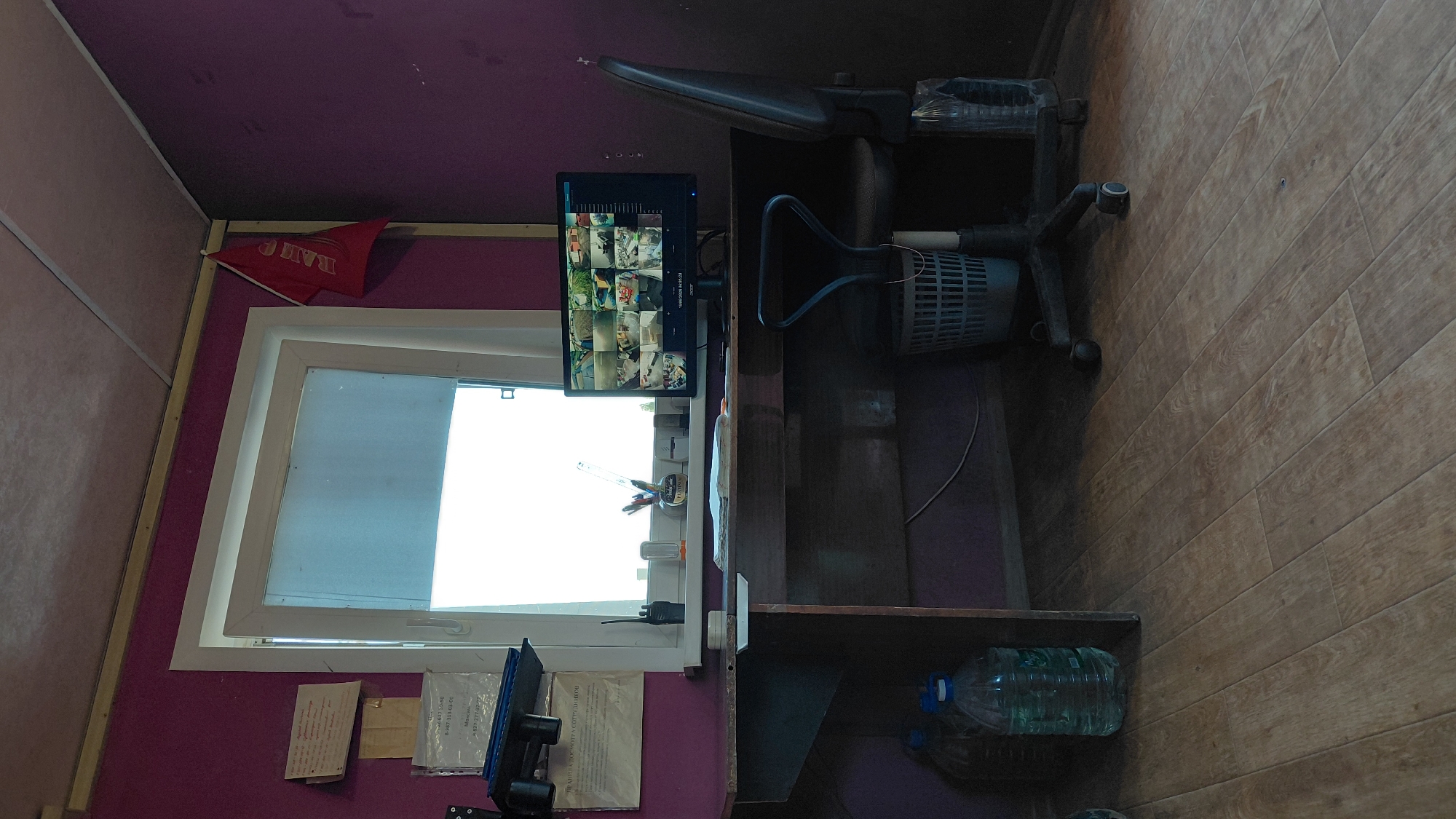 A security monitoring station is depicted in this image. The room features purple walls and a wooden floor. A wooden desk is positioned in front of a window. On the desk, a computer monitor displays multiple security camera feeds, alongside a desk organizer with pens and pencils, a radio, and a small bottle labeled "PUDING." A dark office chair with wheels is situated at the desk, with a trash can beneath it. Two large plastic bottles of liquid are visible on the floor to the left of the desk. A red flag with Cyrillic lettering is mounted on the wall near the window. No individuals are present in the scene. The image likely depicts a security post or control room in Saratov, Russia, given the Cyrillic writing.