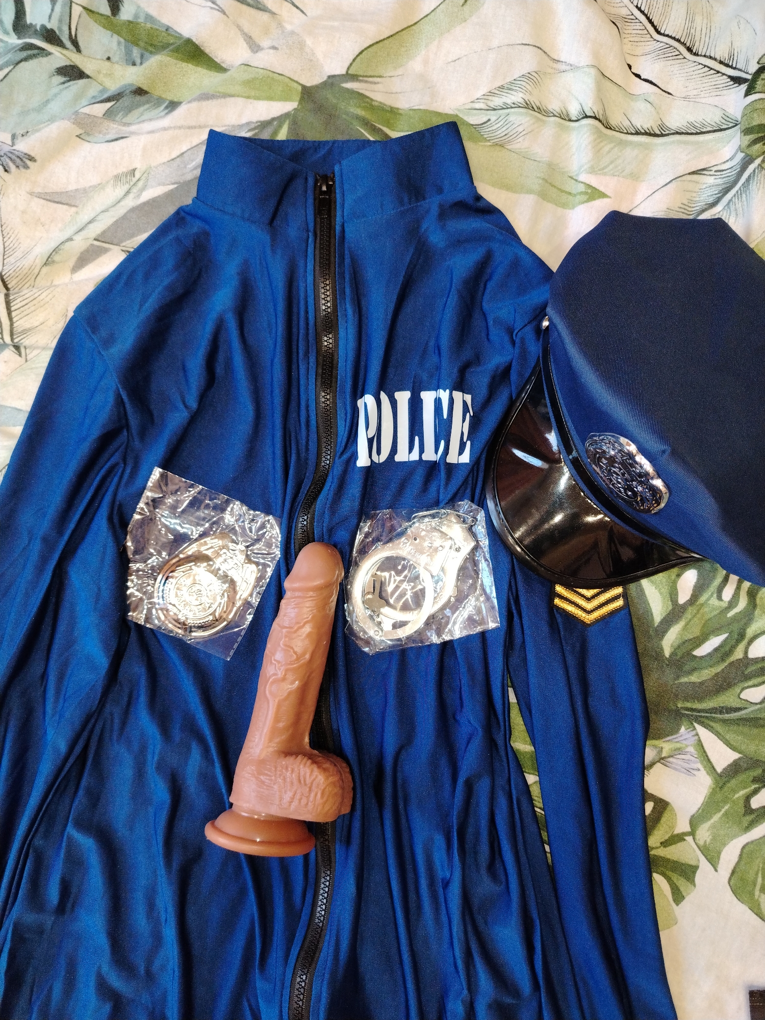 The image shows a police costume laid out on a bed with a white and green patterned bedspread. The costume consists of a blue zip-up jacket with the word "POLICE" printed in white across the chest, a police hat with a shiny black bill and badge, a pair of packaged handcuffs, a packaged badge, and a brown dildo. The location is not visible.
