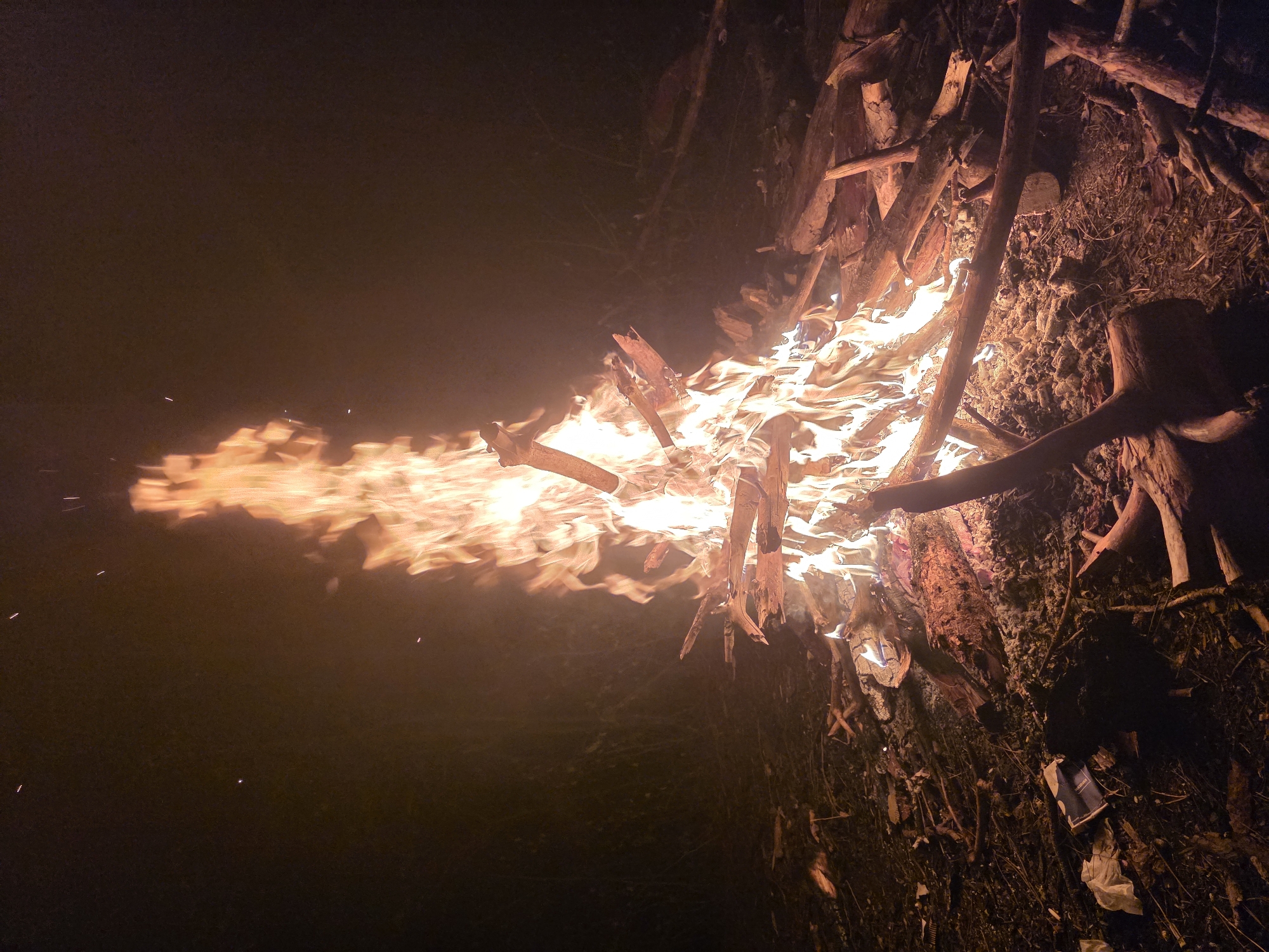 The image shows a bonfire at night. The bonfire is comprised of wooden logs and branches, arranged to create a teepee-like structure that is actively burning. The flames are intense and extend outward, casting a bright orange glow. The surrounding area is dark, with visibility limited to the immediate vicinity of the fire. Details of the surrounding environment are difficult to discern due to the darkness, but it appears to be an outdoor setting with visible ground cover and scattered debris around the base of the fire. There are no visible people in the image. The location is Unknown, Unknown.