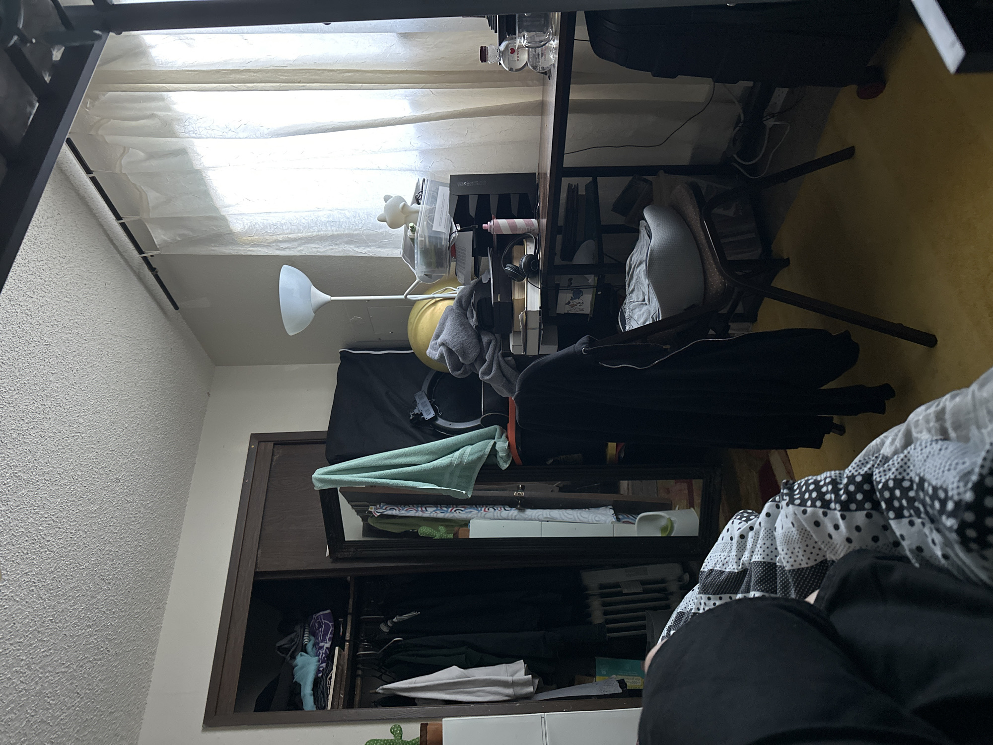 The image depicts a cluttered bedroom. A closet filled with clothes is visible on the left. In the center, a desk is piled with various items including papers, a lamp, headphones, and a pink spray can. A chair with a pillow on it sits in front of the desk. A window with sheer curtains is on the right, letting in natural light. The floor is covered with a yellow carpet. The overall impression is of a lived-in, disorganized space. The location is unknown.