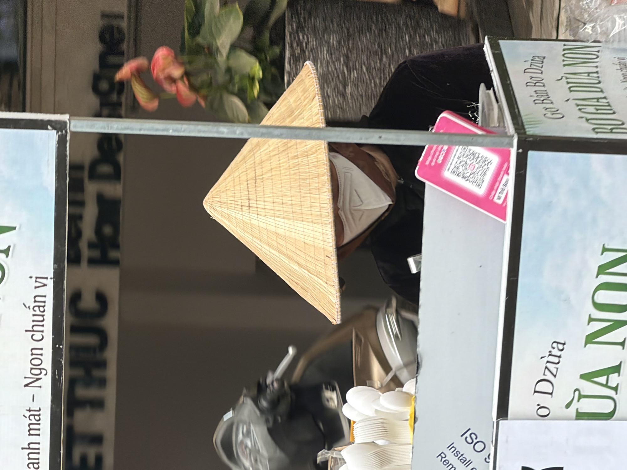 The image depicts a street vendor in Nha Trang, Vietnam, wearing a traditional conical hat and a face mask. The vendor is standing behind a stall, which displays various signs and products. A scooter is visible in the background, partially obscured. The stall itself has a variety of items, including stacks of white disposable cups or utensils, and a pink card with a QR code. One sign on the stall reads "NGON CHUAN VI" and another appears to say "GƠ BÁN BƠ DỪA BƠ GIÀ DỪA NON". The vendor is likely selling food or drinks. The overall scene suggests a typical urban street scene in Vietnam, with a focus on commercial activity.