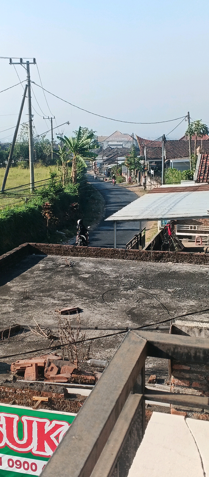 This is a medium-angle, eye-level, outdoor, daytime shot taken in Bocek, Indonesia. The scene depicts a rural road winding through a village. In the foreground, a flat, weathered rooftop is visible, made of concrete. Some debris, including red bricks and dry vegetation, is scattered on the rooftop. A metal railing runs along the edge of the rooftop in the lower right quadrant of the image.
Beyond the rooftop, the road slopes upwards and curves to the right. A person wearing a black jacket and helmet rides a dark-colored motorcycle in the foreground on the road, moving away from the camera. Further up the road, there are several other people and motorcycles, suggesting moderate activity.
The road is flanked by houses with traditional tiled roofs on the right side, and a grassy field on the left. Banana trees and other lush green vegetation line the left side of the road. Utility poles with numerous wires crisscross the sky above the scene. In the background, more houses and the hazy outline of distant hills can be seen. The sky is clear and pale blue, indicating a bright, sunny day. A green and white banner with red lettering is partially visible in the lower left corner of the frame, displaying the text "SUK" and a phone number "1 0900".