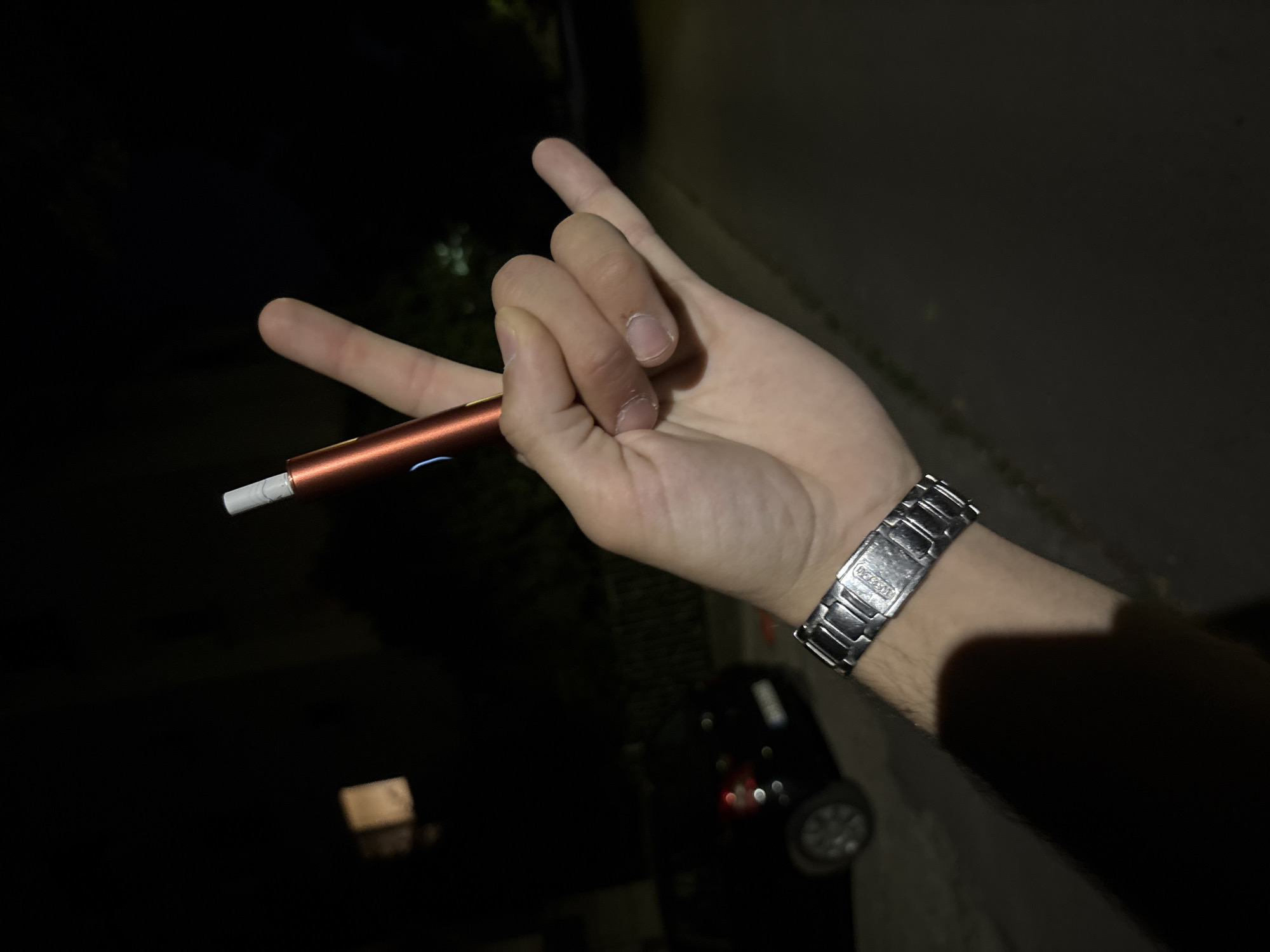 A hand with a metallic watch on the wrist is shown in the foreground, holding a red electronic cigarette with a white tip. The hand is making the "shaka" or "hang loose" gesture. In the background, a dark-colored car is parked. The scene appears to be outdoors at night, with some artificial lighting illuminating the surroundings. The image was taken on the street in front of the residential building located at **ul. "Voynishko Shatra" 68, 1680 Sofia, Bulgaria**.