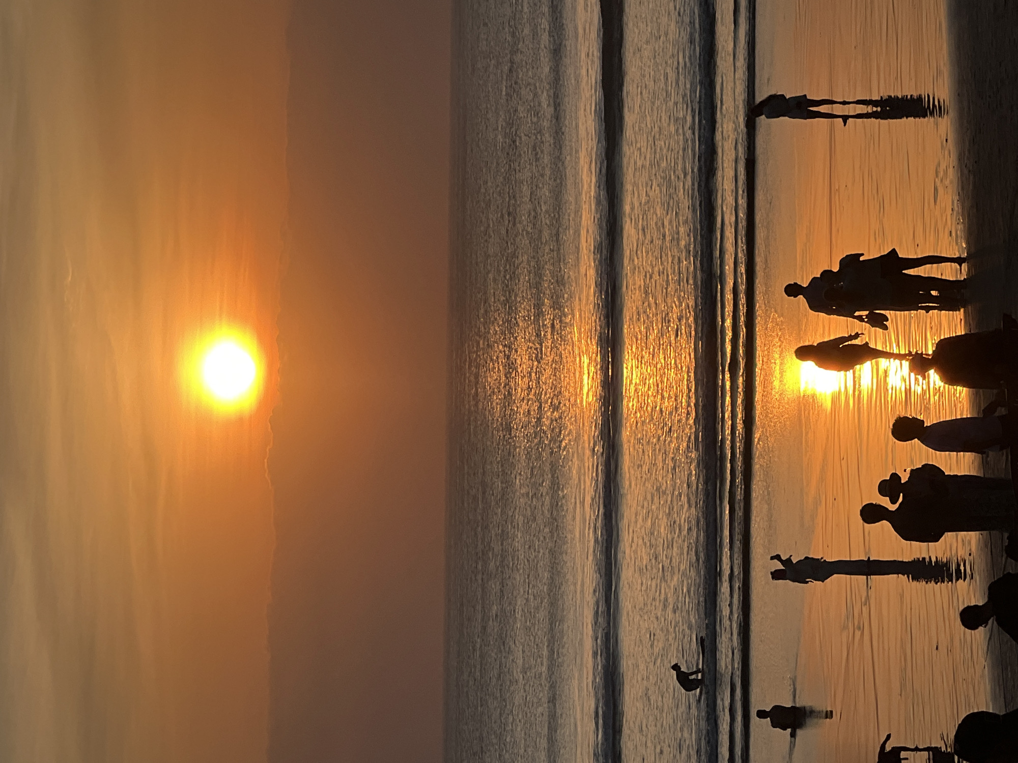 This image depicts a sunset scene at Kuta Beach, Bali, Indonesia. Silhouettes of people are visible on the wet sand and in the shallow water. The sun, a bright orb, is low on the horizon, casting a warm, orange glow across the sky and reflecting on the water. Waves are breaking gently on the shore. Several individuals are standing and walking, some appearing to take photos or videos, while others are interacting. In the water, one person is on a surfboard. The overall atmosphere is peaceful and serene, characteristic of a beach sunset.