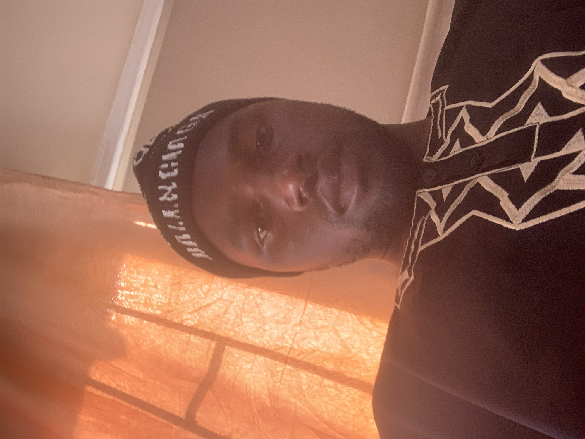 In Gura Top, Nigeria, an overhead medium shot shows a man wearing a black beanie with white lettering and a black garment with intricate white embroidery. The man is facing the camera directly, with his head tilted slightly. The background features a sheer orange curtain with light filtering through it, creating a warm, diffused glow. To the right of the curtain, a plain beige wall is visible. The overall lighting is soft and ambient.