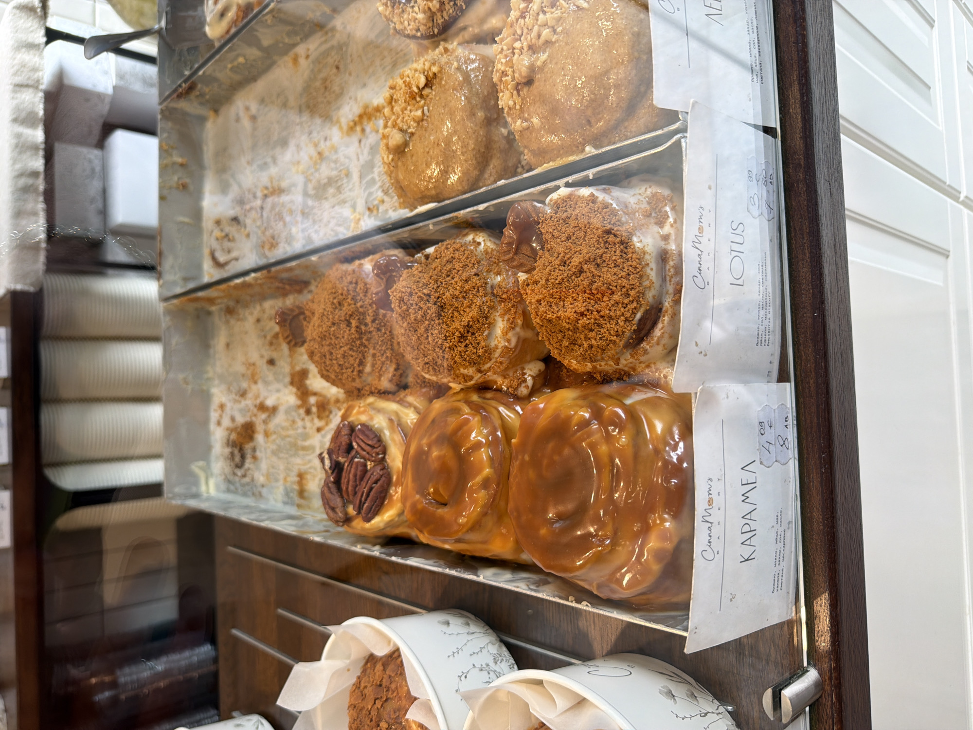 The image depicts a pastry display case with various types of cinnamon rolls or sweet buns at "Cinna Mom's" in Sofia, Bulgaria. There are three shelves visible, each containing a different flavor or topping. The top shelf has rolls covered in a light brown topping with nuts. The middle shelf features rolls with a crumbly, dark brown topping and a small chocolate swirl. The bottom shelf displays rolls coated in a shiny caramel glaze, with some featuring pecan halves. Small white paper cups containing individual pastries are visible at the bottom of the case. There are price labels attached to the display case, indicating the names of the pastries as "LOTUS" and "КАРАМЕЛ" (Caramel) with their respective prices in Bulgarian currency (лв).