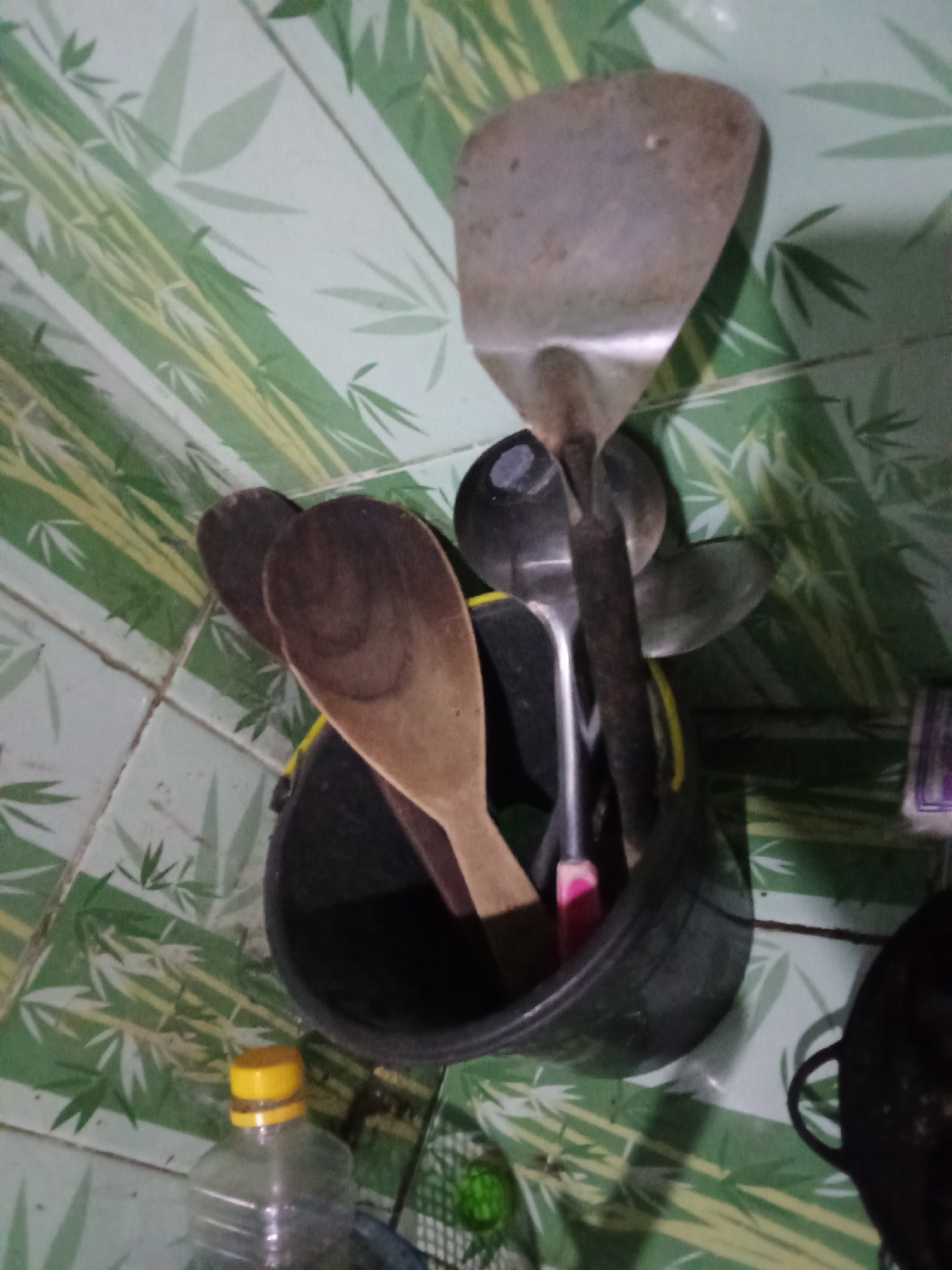 A still life of kitchen utensils in a bucket is displayed against a backdrop of green bamboo-patterned tiles. In the foreground, a partially filled plastic bottle with a yellow cap sits on the floor. Behind it, a black bucket contains various cooking implements. A large, worn metal spatula with a wide, flat head rests at the top, angled slightly to the right. Below it, a smaller metal spatula or ladle is visible. Two wooden spoons, one with a darker, more pronounced grain, are also prominent in the bucket. The overall scene suggests a utilitarian and perhaps well-used kitchen environment, likely within a residential dwelling in Pemalang, Indonesia. The lighting is dim, casting shadows and highlighting the textures of the objects.