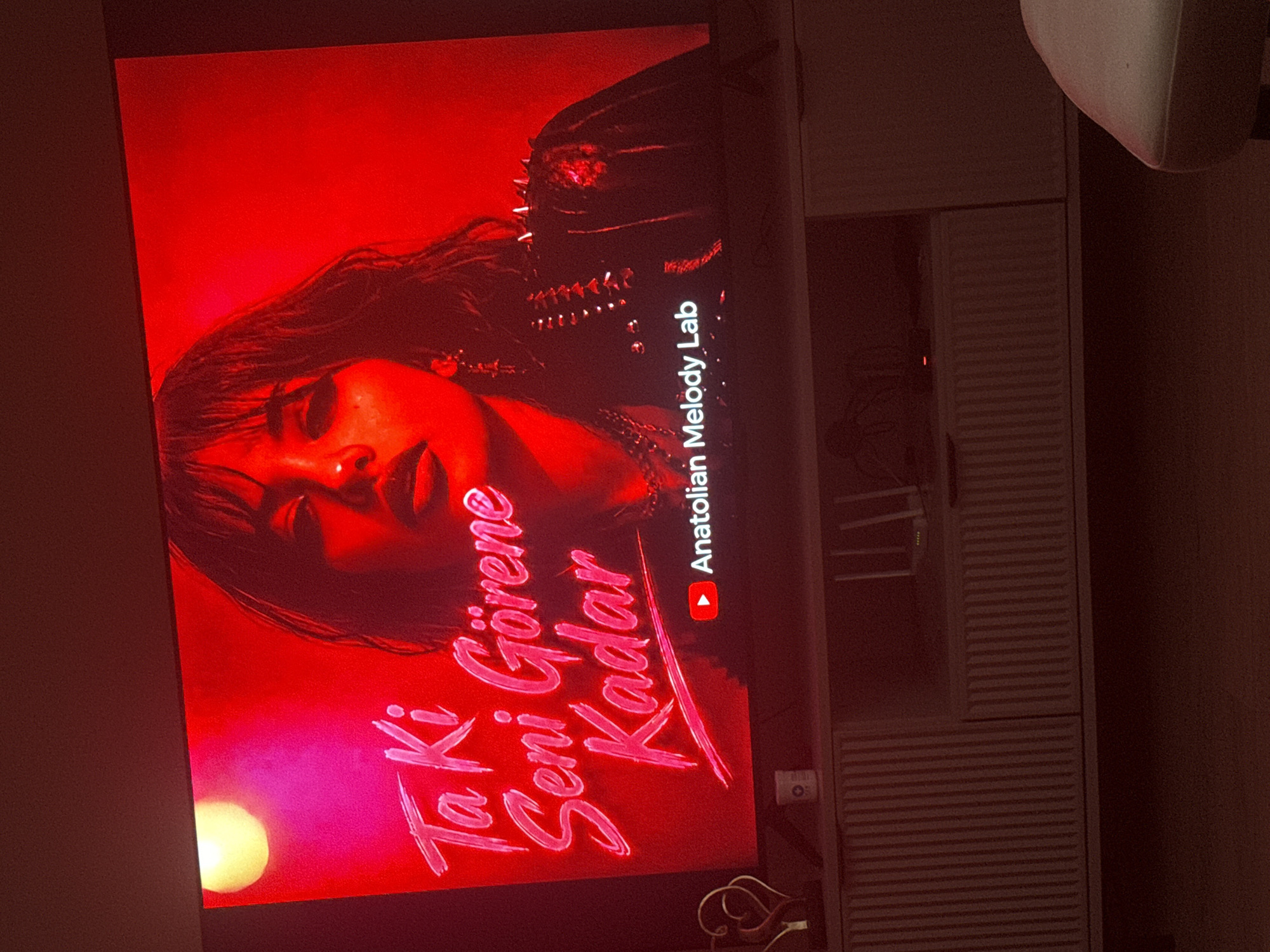 The image shows a television screen displaying a red-toned music video by "Anatolian Melody Lab" titled "Ta Ki Seni Görene Kadar." A woman with dark hair and dark clothing is visible in the video. The TV is positioned in what appears to be a home entertainment setup, with a white storage unit visible to the right of the television.