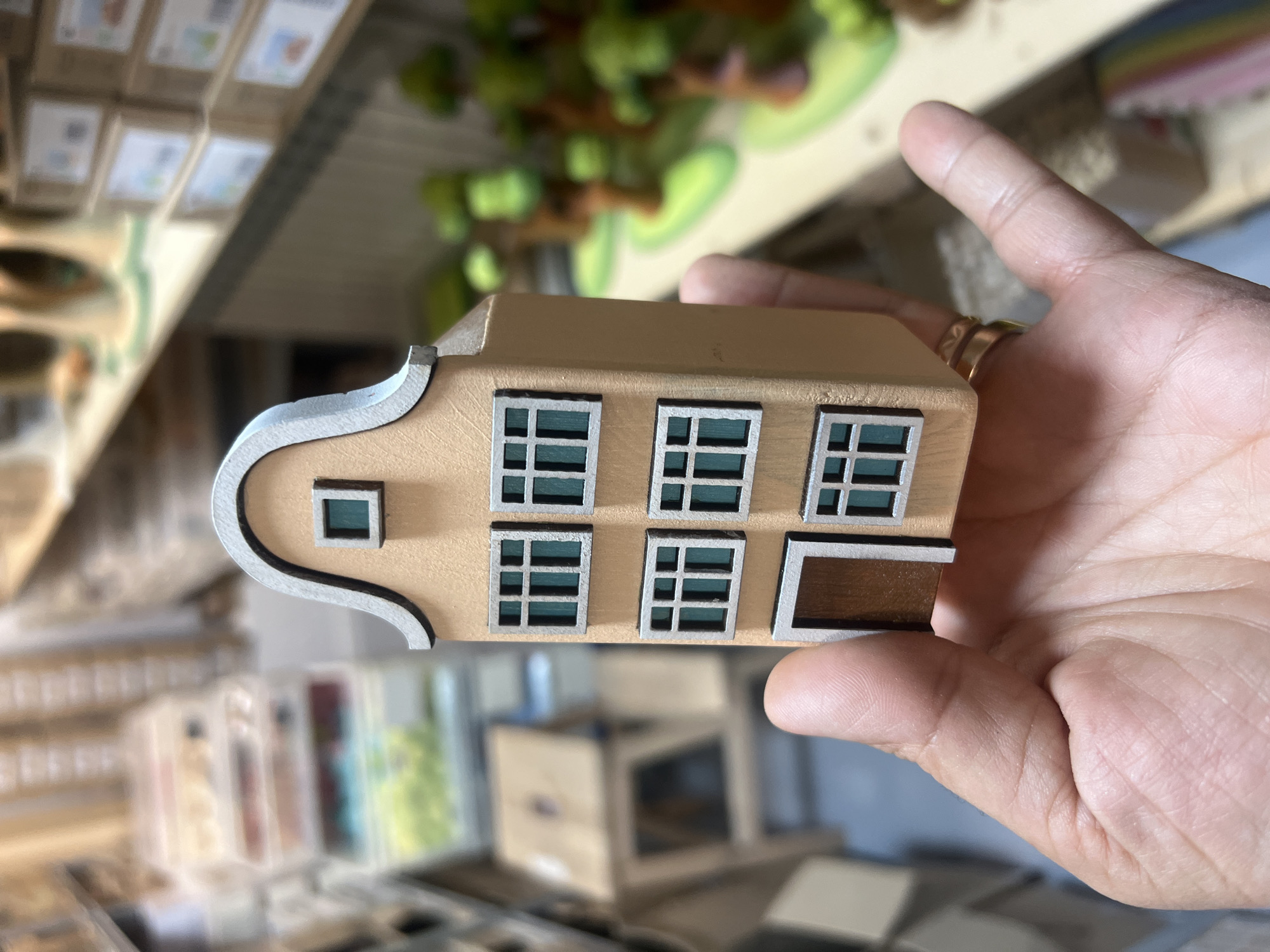 A close-up shot shows a hand holding a miniature model of a multi-story building. The building model is a light peach color with a grey curved roof. It has several windows, each divided into four panes, and a single brown door. The hand, with visible lines and a ring on the finger, supports the model. In the blurred background, shelves are stocked with various items, including what appear to be boxes and other small decorative objects, suggesting a retail or craft environment.
