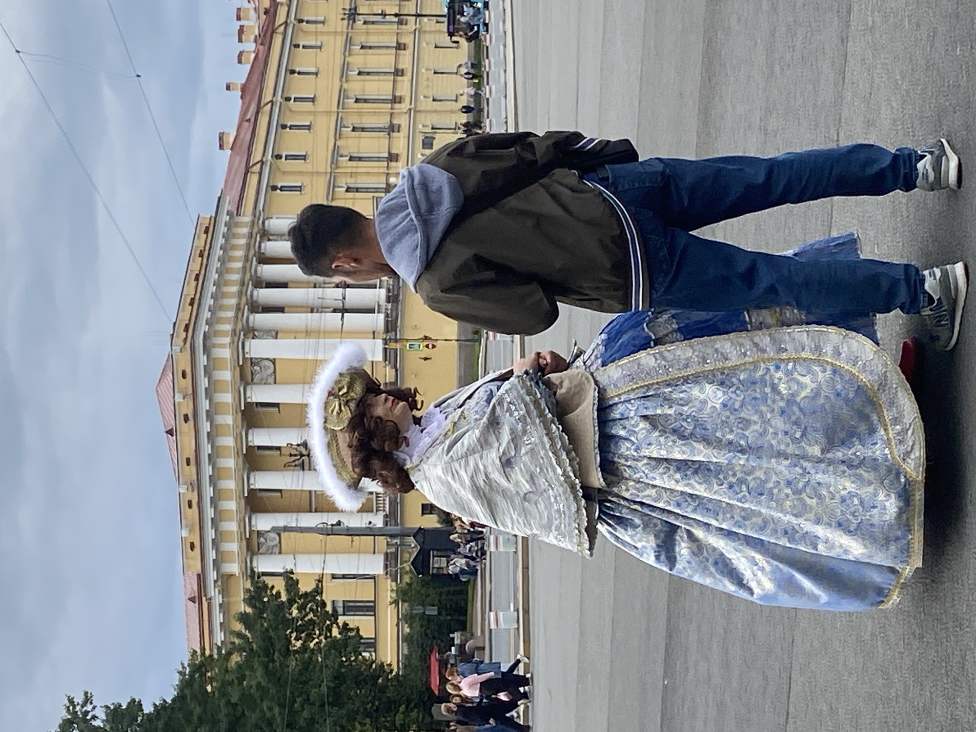 In Saint Petersburg, Russia, on an overcast day, a person dressed in a historical costume stands in a public square, facing another person who is wearing modern clothing. The person in the historical costume is adorned in a blue and white patterned gown, a white cape with intricate silver detailing, and an elaborate hat with feathers and gold embellishments. Their hair is styled in curls, and they are wearing a ruffled collar. They are looking to their left, towards the person in modern attire.
The second individual is a young man wearing a dark green jacket with a grey hood underneath, blue jeans, and grey sneakers. He is facing the person in the costume, with his hands clasped in front of him. His gaze is directed towards the costumed individual.
Behind them, a grand yellow building with white columns and a red roof dominates the background. The building appears to be an important architectural landmark. In the middle ground, there are other people walking, some with shopping bags, and a bus can be seen on the right side of the image. The ground is paved, suggesting an urban setting. The sky is cloudy, casting a diffused light on the scene. The image appears to be taken on Palace Square, a prominent public space in Saint Petersburg known for its historical significance and architectural grandeur.