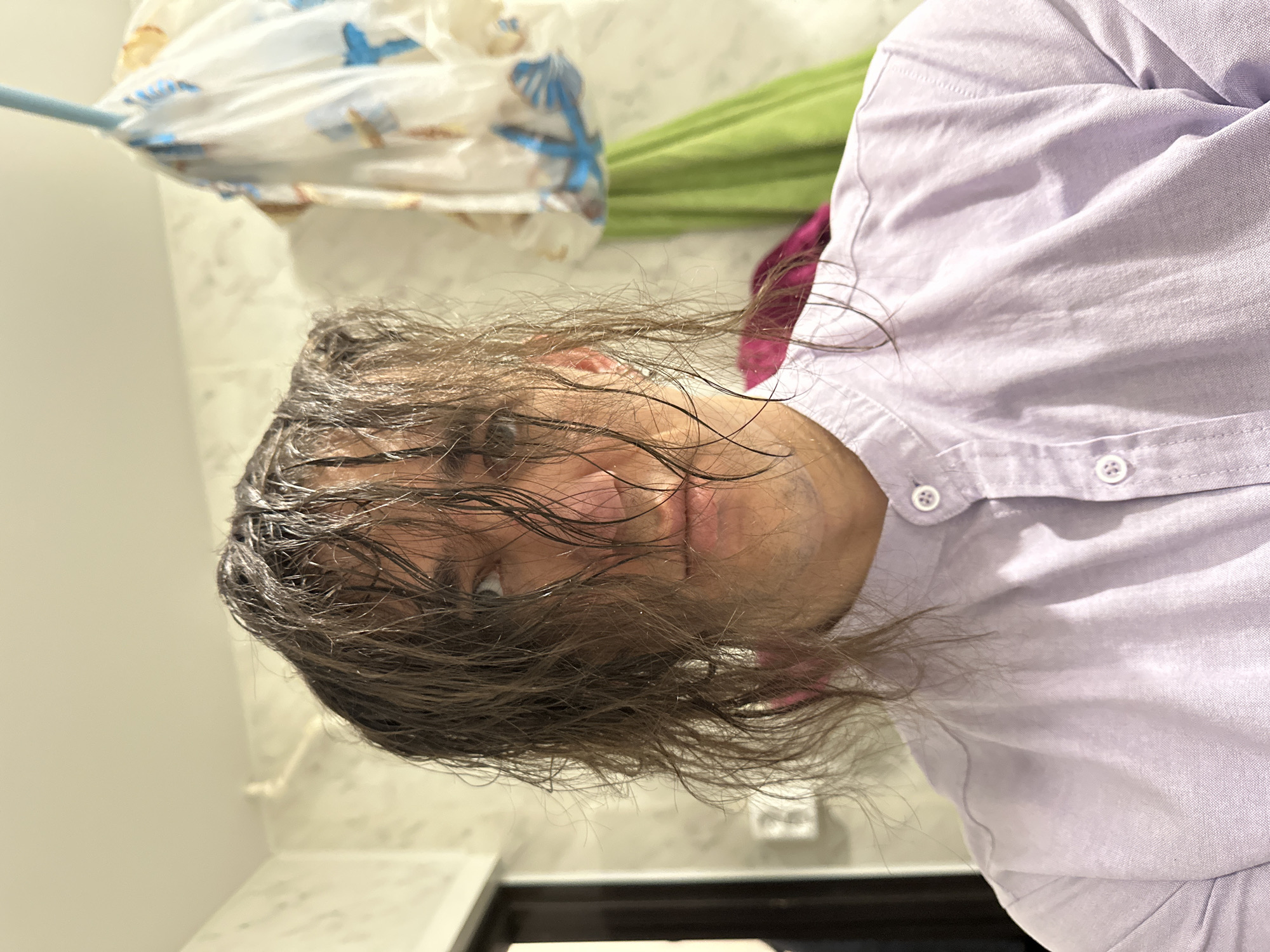 A close-up, portrait-style photograph shows a man with wet, dark brown hair and eyes looking directly at the camera. He is wearing a light purple, collared shirt with white buttons. Behind him, blurred, is a white shower curtain with blue and green designs. A green object is also visible. The background suggests a bathroom setting. There is no evidence to suggest the location is Saratov, Russia.