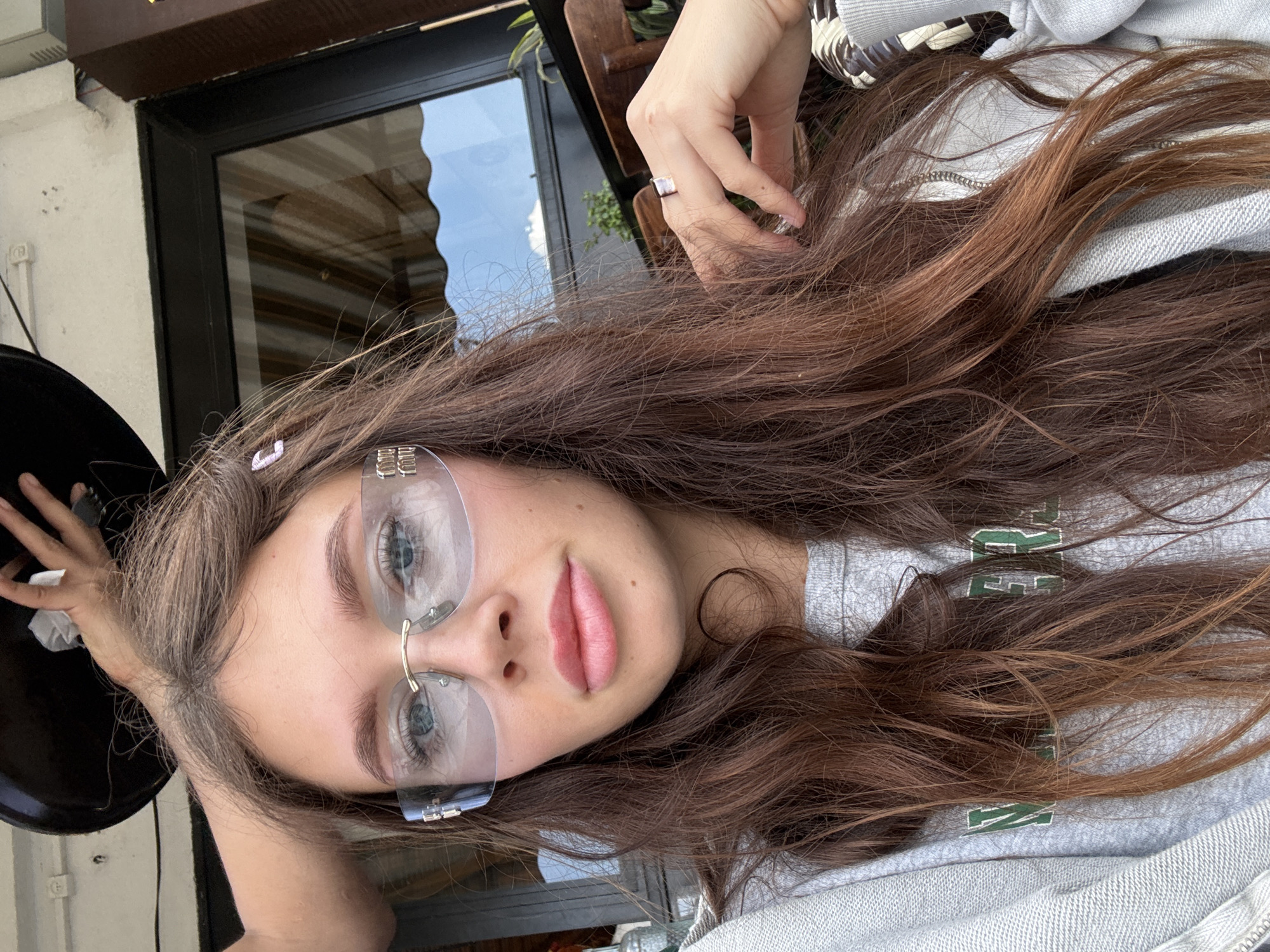 The image depicts a young woman with brown hair and blue eyes, wearing rimless glasses. She appears to be sitting at an outdoor cafe in an unknown location, as evidenced by the partially visible window, greenery, and table with chairs in the background. She is wearing a gray sweatshirt with green lettering. Her hand is raised near her face, and she is wearing a ring on her finger. The photo is taken from a close-up, slightly angled perspective.