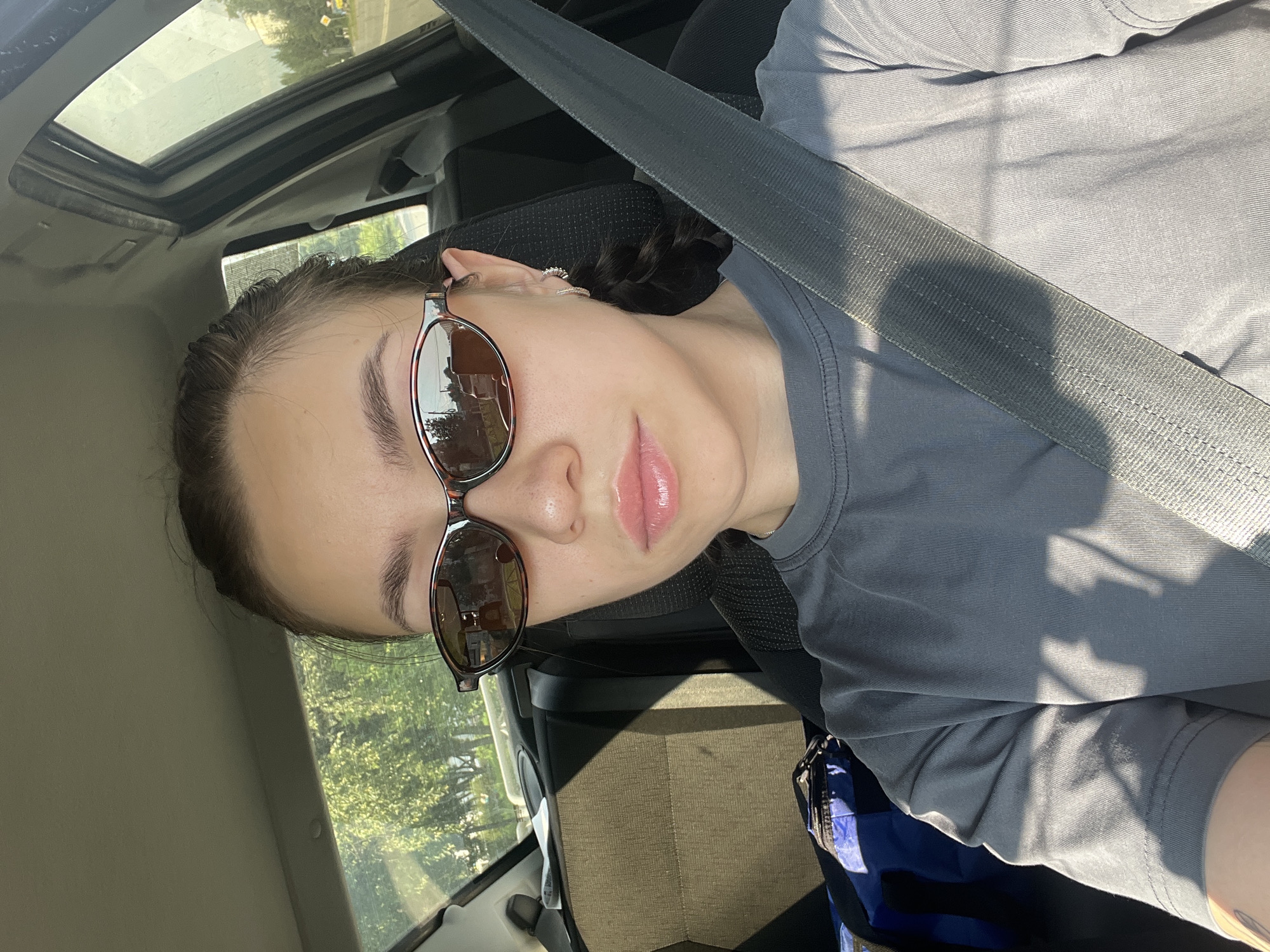 A young woman is sitting in the driver's seat of a car, looking directly at the camera. She is wearing sunglasses and a grey t-shirt. The car interior is visible behind her, including the seatbelt across her chest, the car seat, and the headrest. Sunlight streams through the windows, casting shadows. Outside the car, trees and a road are visible, suggesting an outdoor, possibly suburban or rural, setting. The image is taken from a selfie perspective.