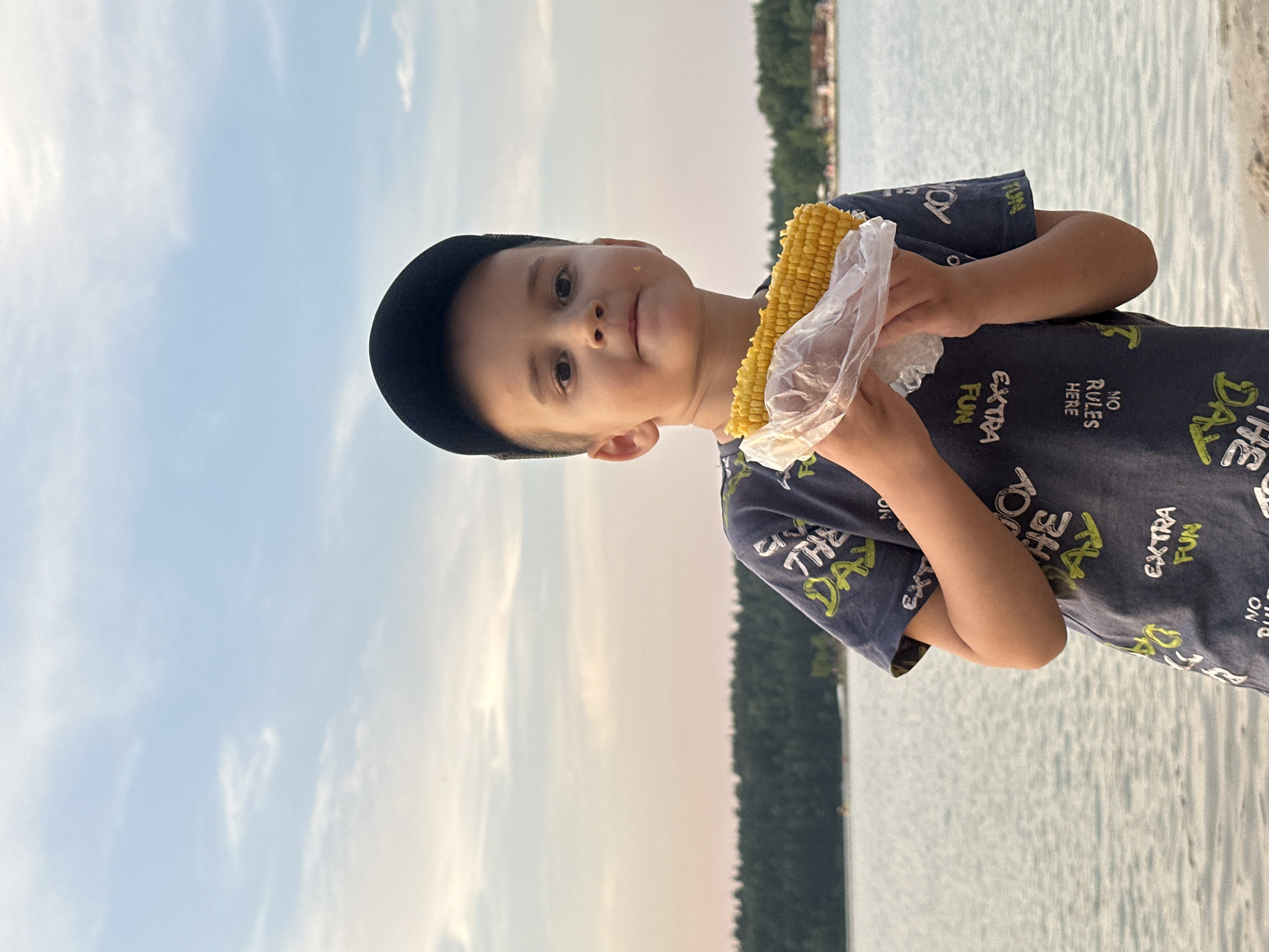 A young boy wearing a black baseball cap and a dark t-shirt with a pattern of text and graphics stands on a sandy shore, holding a cob of corn wrapped in plastic. He is looking at the camera with a slight smile. The background shows a calm body of water, possibly the Oka River, with a line of trees in the distance. The sky is partly cloudy with soft, muted colors, suggesting it is either sunrise or sunset. The overall scene conveys a casual, outdoor setting. The location is Lytkarino, Russia.