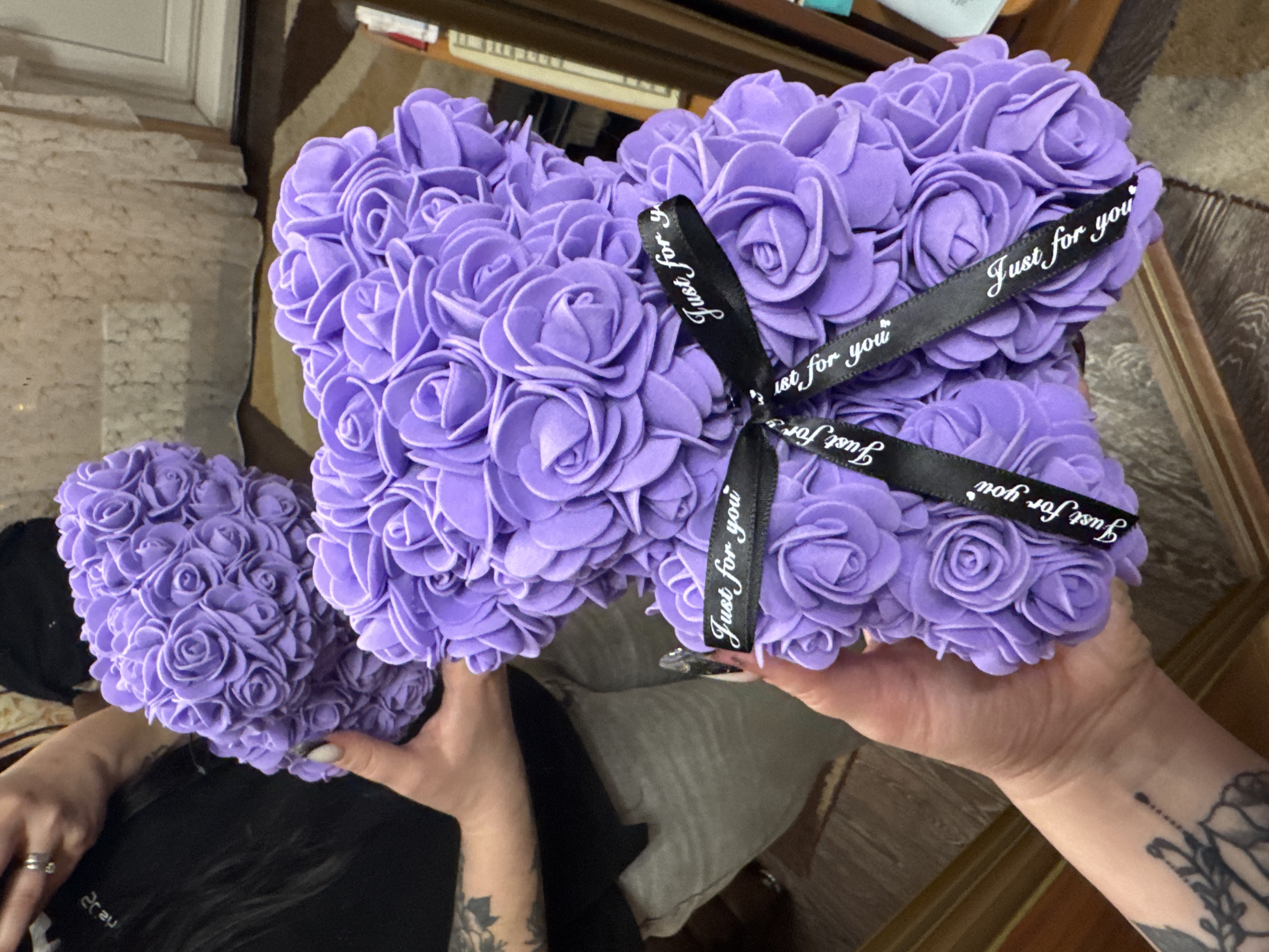 Here is a detailed description of the image:
Two purple rose-covered objects are held by a person with tattoos on their arms. The object in the person's right hand is larger and has a black ribbon wrapped around it with white lettering that says "Just for you." The object in the left hand is smaller and rounder. The background appears to be an indoor setting.