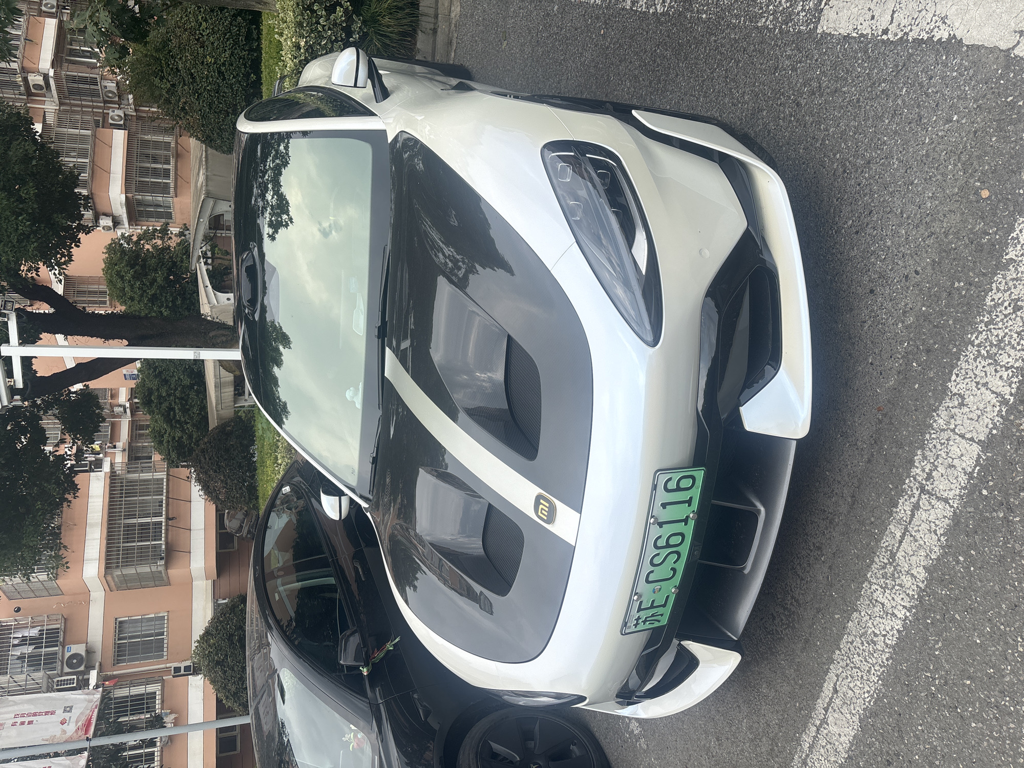 The image shows a white sports car parked on an asphalt road. The car has a black hood with two white racing stripes and a black front bumper. A green license plate with the characters "E-CS6116" is visible on the front bumper. The car is parked next to a black car, and in the background, there is a building with pink and white walls, and trees. The scene appears to be in a residential area of Suzhou Industrial Park, China, given the presence of apartment buildings common in that region and the license plate format.