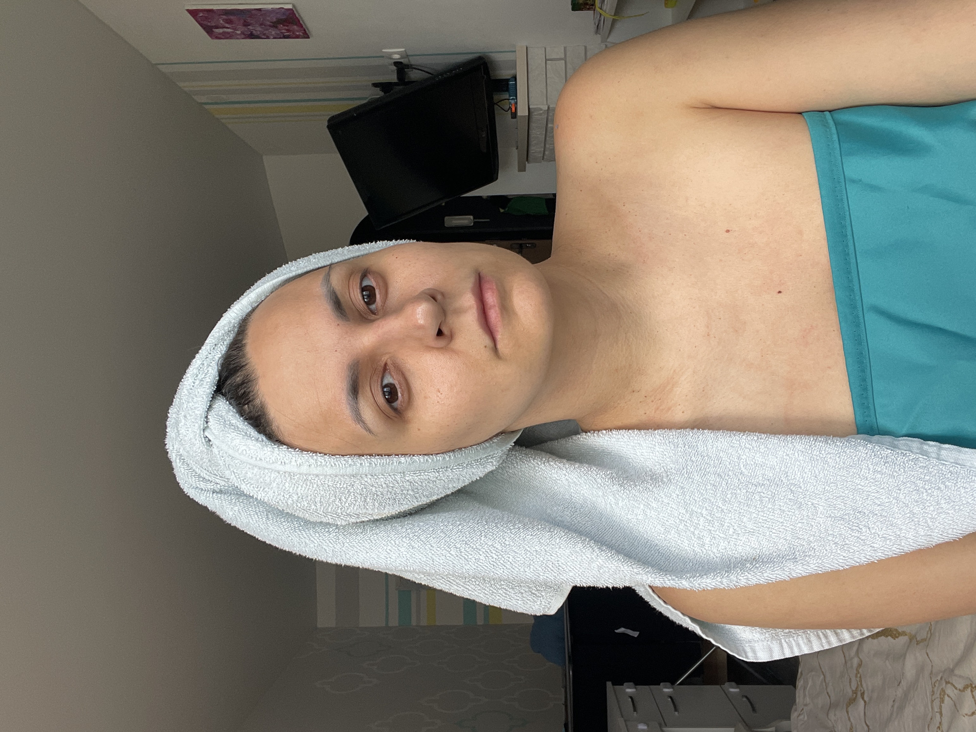 A medium shot shows a woman with a light blue towel wrapped around her head and chest, standing in what appears to be a bedroom. She has light skin, brown eyes, and her eyebrows are lightened. She is looking directly at the camera with a neutral expression. Her shoulders and upper chest are visible, and she is wearing a turquoise bandeau. The background includes a wall with striped wallpaper, a television set on a shelf, and a dresser with drawers. The room is lit by natural light coming from the right side of the image. The overall impression is that of a person who has just showered or is preparing for bed. The location in Izhevsk, Russia is not identifiable from the image itself.