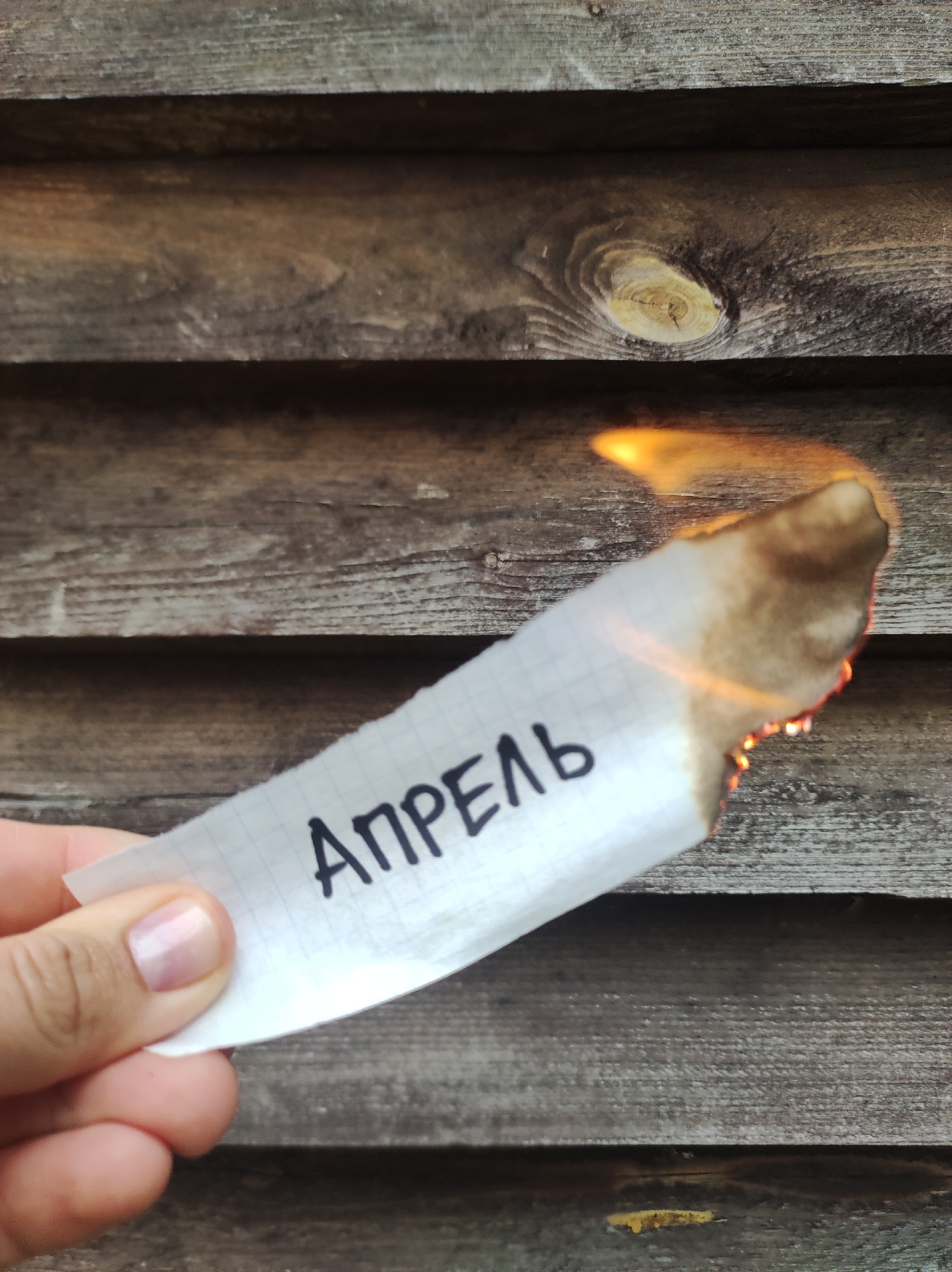 A photograph shows a person holding a piece of paper with the Russian word "АПРЕЛЬ" (April) written on it, with the edge of the paper on fire. The background is a wooden wall with horizontal planks. The photo appears to be taken outdoors. There is no information indicating the location in the city of Фоки, Russia.