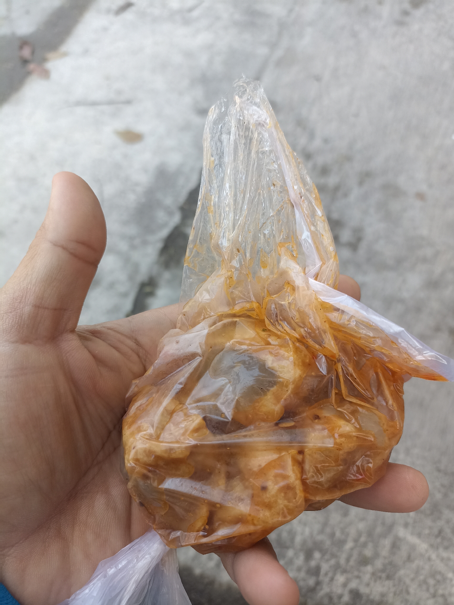 An overhead, close-up shot shows a hand holding a clear plastic bag of food. The food appears to be small, fried pieces coated in a reddish-orange sauce. The plastic bag is slightly crinkled and has some of the sauce on the outside. The hand is a light-skinned adult hand, with the thumb and fingers gently cupping the bag. The background is a light gray, textured surface, possibly concrete, which could be a sidewalk or pavement. The lighting is bright and natural, casting soft shadows. There are no visible identifying landmarks or signs of the city of Wonokromo, Indonesia, in this image.