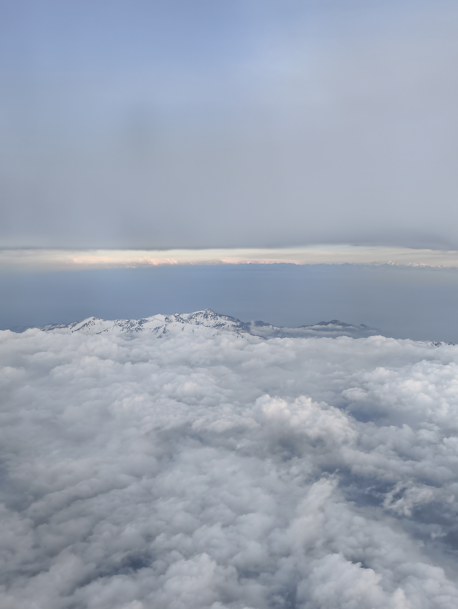 The image shows a mountain range with snow-capped peaks jutting out above a dense layer of clouds. Above the mountains, there is a band of lighter, possibly sunlit clouds, followed by a grey sky. The location is in the city of Unknown, Unknown.