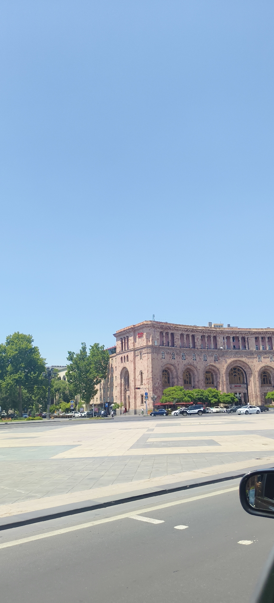 A wide shot captures a sunny day in Yerevan, Armenia, with a clear blue sky dominating the upper half of the frame. In the mid-ground, a grand, multi-story building constructed of pinkish stone stands prominently. Its facade features numerous arched windows and classical columns. A red sign with white lettering is visible near the top of the building.
To the left of the stone building, several large, leafy green trees provide shade, and behind them, other buildings of varying architectural styles can be seen. A wide, paved square occupies the foreground and extends towards the buildings. The square is made of large, light-colored tiles with some darker accents forming patterns.
A road runs along the edge of the square. Several cars are parked and driving on the road, including white, black, and blue vehicles. Pedestrians are walking on the sidewalks and in the square. The image is taken from a slightly elevated position, possibly from a vehicle, as a car's side mirror is visible in the lower right corner, reflecting parts of the scene. The overall impression is of a well-maintained urban space on a bright day.