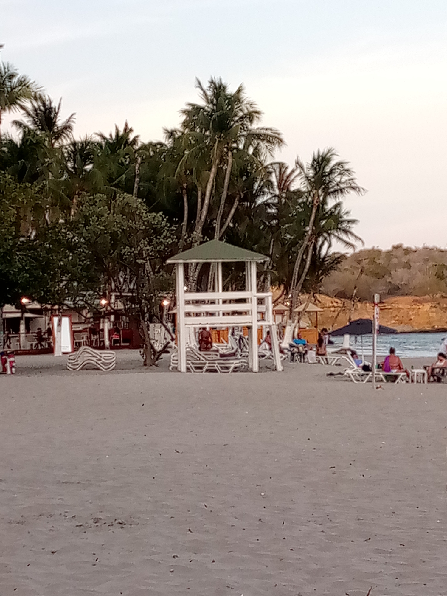 The image shows a beach scene in El Yaque, Venezuela. In the foreground is a wide expanse of sandy beach. In the midground is a white wooden lifeguard tower with a green roof. Several people are scattered around the beach, some sitting on lounge chairs under umbrellas. Behind the lifeguard tower, there are palm trees and other greenery. To the left of the lifeguard tower is a building with lights, possibly a restaurant or bar. In the background is a slightly elevated, brownish landscape. The sky is a pale, muted color, suggesting dusk or dawn.
