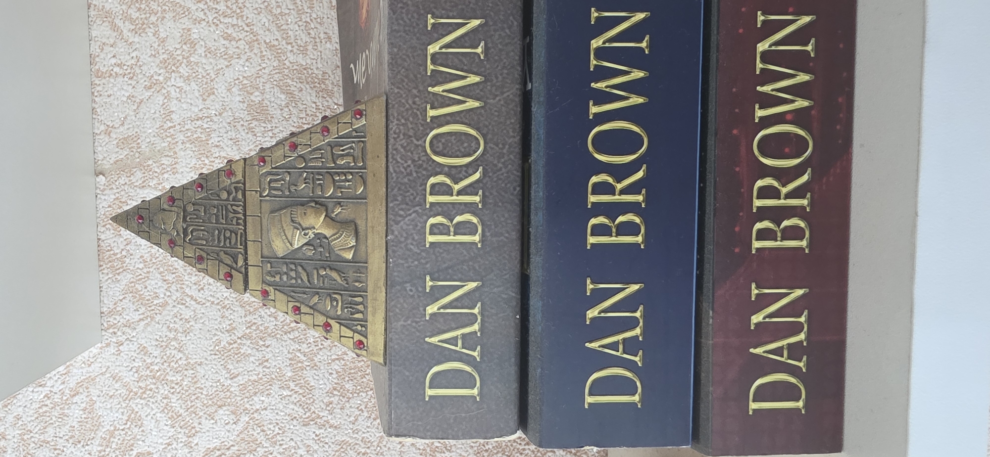 The image shows a close-up, high-angle view of three books stacked vertically. The top book has a dark gray cover with gold lettering that reads "DAN BROWN". It is partially obscured by a decorative pyramid with Egyptian motifs and red gem accents. The second book from the top has a dark blue cover, also with gold lettering that reads "DAN BROWN". The bottom book has a dark red cover with gold lettering that reads "DAN BROWN". The books are resting on a white surface. The background is a textured, light-colored wall. There are no people visible in the image. The scene appears to be indoors, possibly on a shelf or table. The location is not specified as being in the city of Sumgayit, Azerbaijan.
