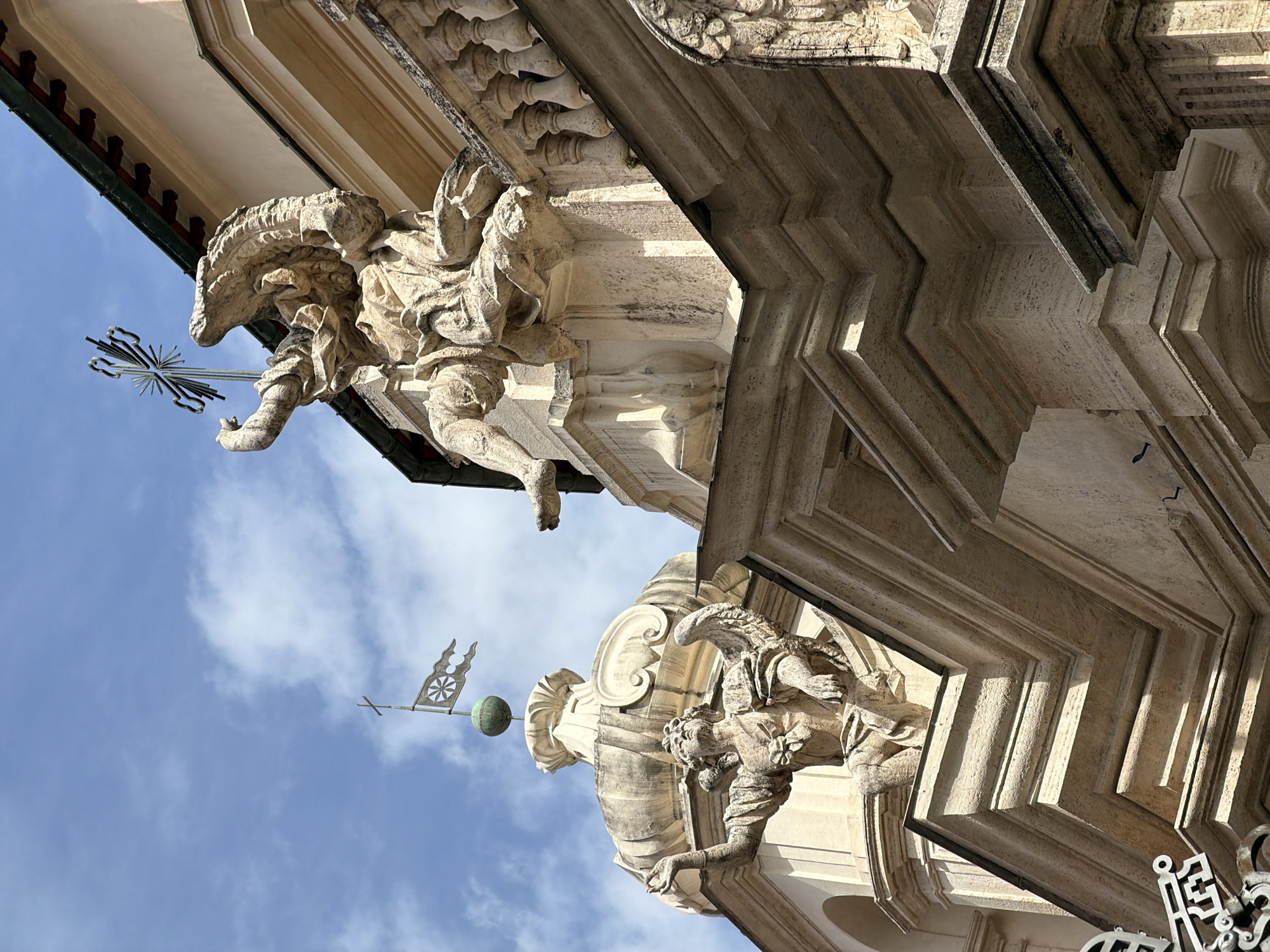 The image presents a dramatic, upward-angled view of an ornate stone building, likely a church or a grand historical structure, set against a bright blue sky with wispy white clouds. The architectural style appears to be Baroque or Renaissance, consistent with its location in Rome, Italy.
Two elaborate stone sculptures are prominent. In the upper left, a dynamic figure, possibly an angel or a mythical creature, is carved into the building's facade. It features an intricate head that resembles a dragon or beast, a human-like torso and legs, and appears to be holding a metal cross with a sunburst motif. The sculpture's form is highly detailed, with visible drapery and muscular definition.
In the lower right, another grotesque or gargoyle-like sculpture with outstretched wings and a detailed, monstrous face extends from the building. Its arm is outstretched, adding to the sculptural complexity.
Between and above the lower right sculpture, a metal finial or weather vane rises towards the sky. It consists of a pole topped with a small cross and a banner-shaped element featuring a spoked wheel design.
The building's stonework is highly decorative, with multiple layers of cornices, moldings, and balusters. The deep, angular cuts and recesses in the facade create striking patterns of light and shadow, emphasizing the texture and three-dimensionality of the architecture. The bright lighting suggests it is daytime. There is no visible text in the image.