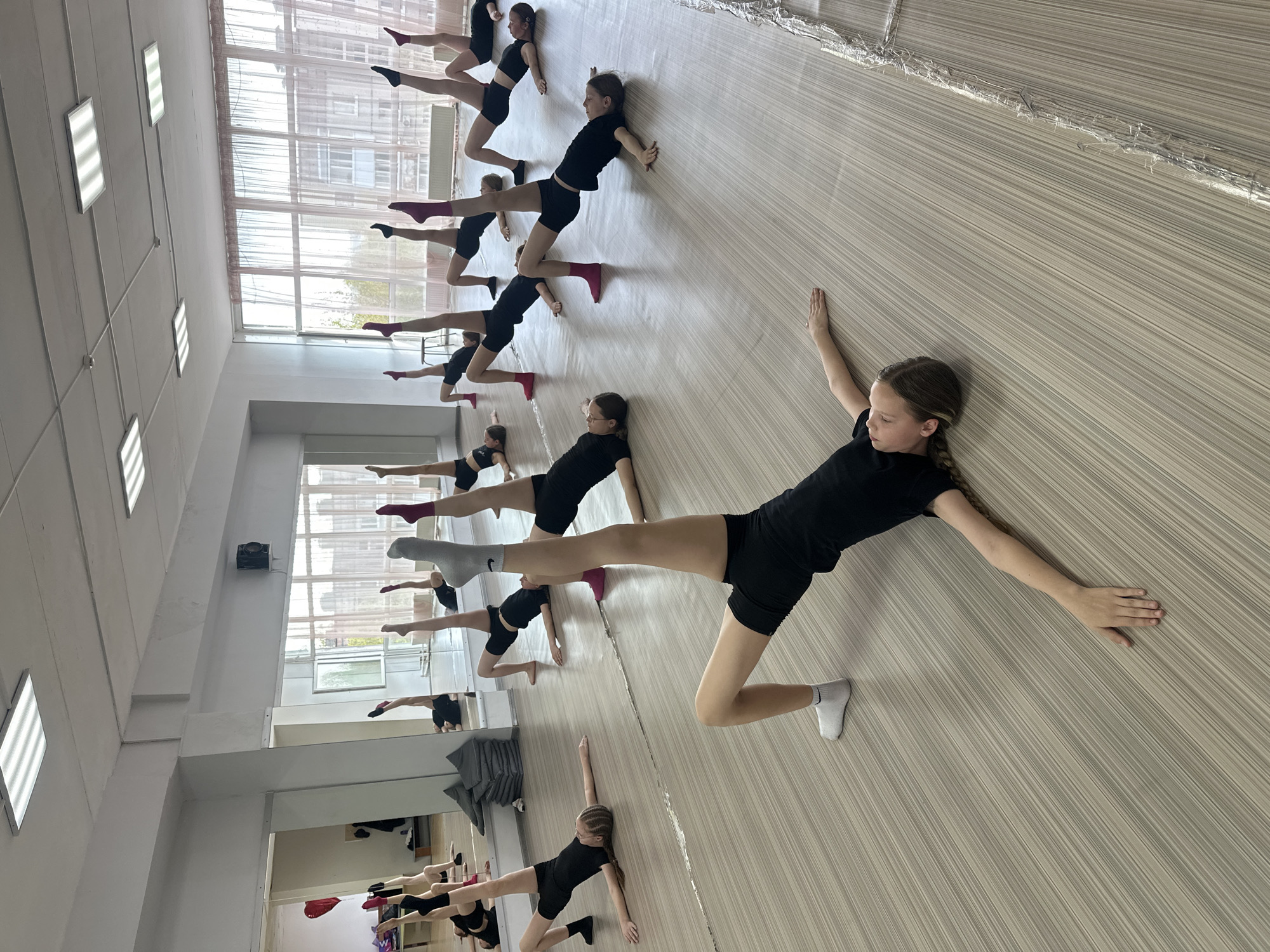 The image shows a group of young girls in a dance studio. They are wearing black tops and shorts and are performing a floor exercise. The girls are arranged in rows across a light-colored, wood-patterned floor. The room has large windows that allow natural light in. The studio appears to be located in Votkinsk, Russia.