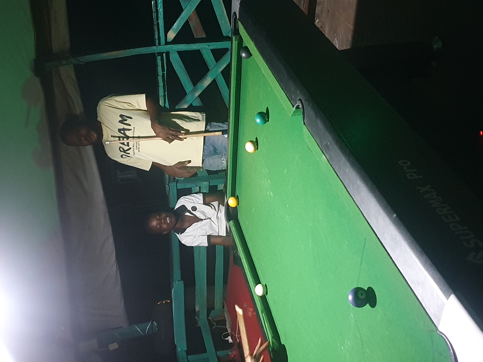 In Lokoja, Nigeria, two individuals are engaged in a game of pool under a covered outdoor area at night. A younger boy in a white polo shirt with a dark collar and a crest on the left chest is seated on a bench behind a green pool table. To his right, an adult male wearing a yellow t-shirt with the word "DREAM" printed in black and blue text, and blue jeans, stands holding a pool cue. The pool table has several balls on it: a white cue ball, a yellow ball, a green ball, a black ball, and a dark-colored ball with a number visible. The environment appears to be rustic, with green wooden railings and a tarp overhead providing shelter. The lighting is artificial, casting shadows and highlighting the pool table. The scene suggests a casual recreational activity.