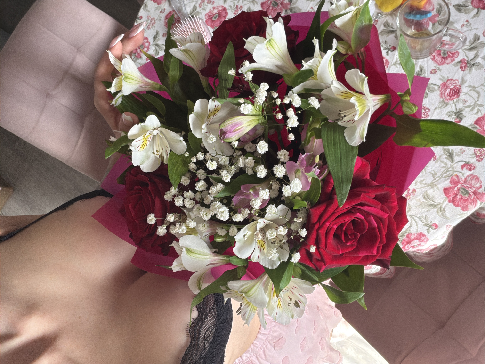 A high-angle, eye-level shot captures a person holding a bouquet of flowers. The bouquet is composed of red roses, white alstroemeria, and baby's breath, wrapped in pink paper. The person's hand is visible, holding the bouquet. A portion of the person's chest is visible, adorned with a black lace garment.
The scene is set indoors. The background includes a patterned tablecloth with floral designs. A glass or mug is visible on the table. The location appears to be in Saint Petersburg, Russia, as the items and arrangements are typical of such settings.