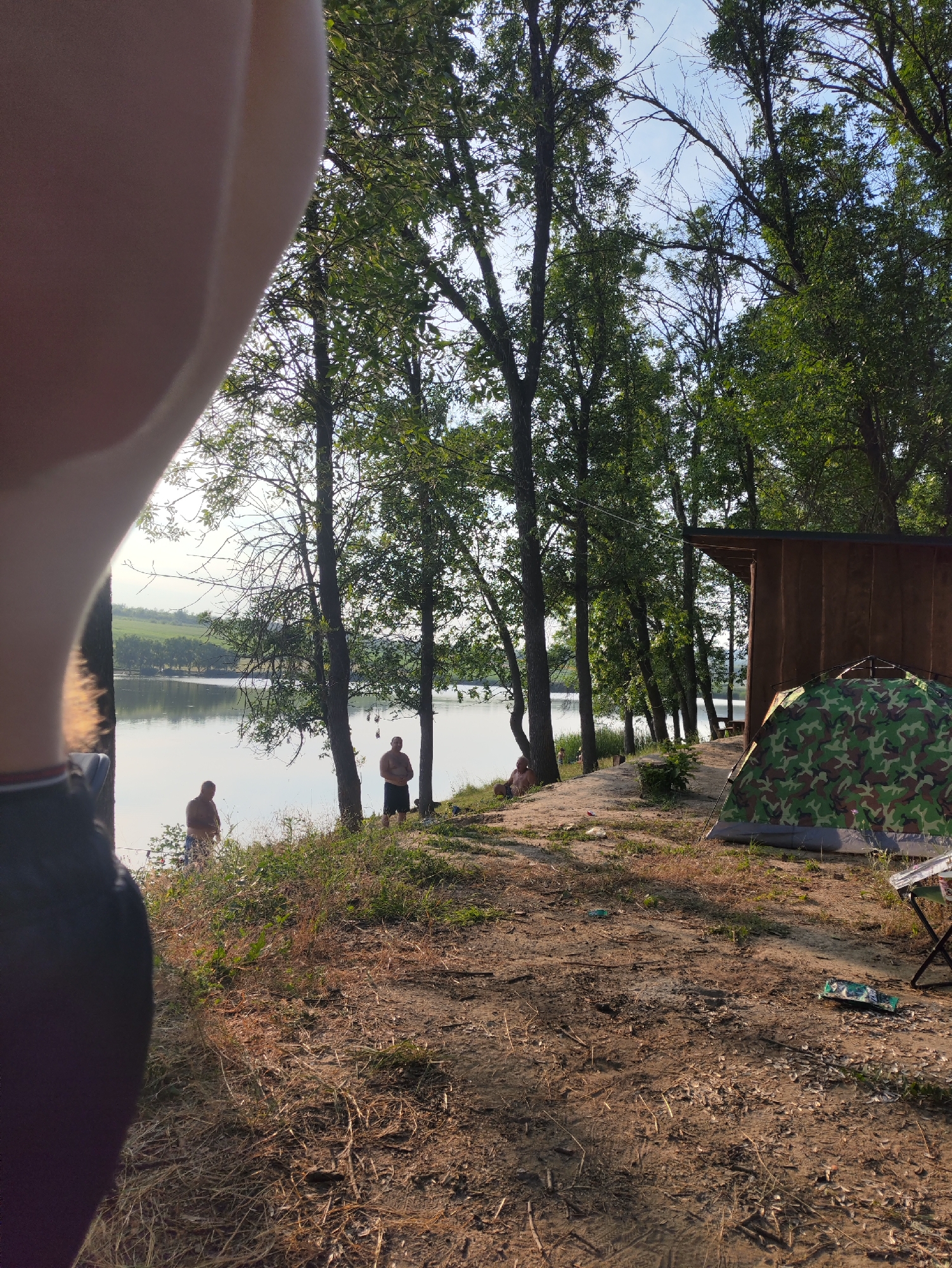 In Găleștii Noi, Moldova, a campsite is set up near a body of water. In the foreground, the left side of the image is dominated by a blurred, out-of-focus section of what appears to be a person's arm or torso. Beyond this, a camouflage tent is pitched on dry, sandy ground littered with twigs and dry leaves. To the right of the tent stands a small, brown wooden structure, possibly a shed or cabin. Several tall trees with green foliage line the background, their branches reaching towards a pale blue sky. Further in the distance, on the shore of the calm water, three individuals are visible. Two men stand near the water's edge, facing away from the camera, while another person sits on the bank to their left. The water reflects the sky and the surrounding trees, creating a peaceful atmosphere.