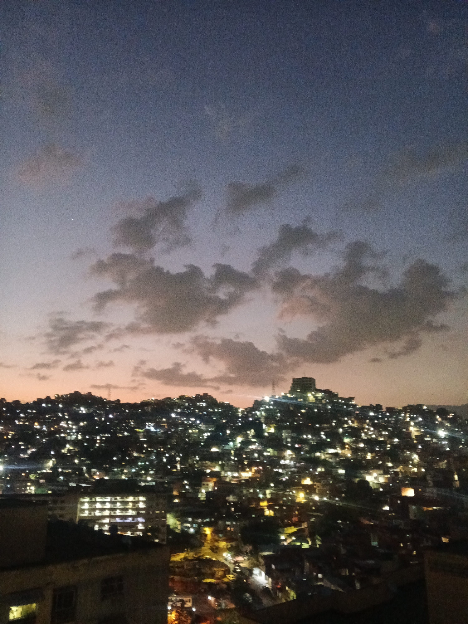 A nighttime view of Caracas, Venezuela is captured under a dusky sky with scattered clouds, their undersides lit by the fading sunset. A city sprawls across rolling hills, densely packed with buildings aglow with numerous lights, creating a stark contrast to the darkening sky. The foreground includes parts of a multi-story building, with illuminated windows.