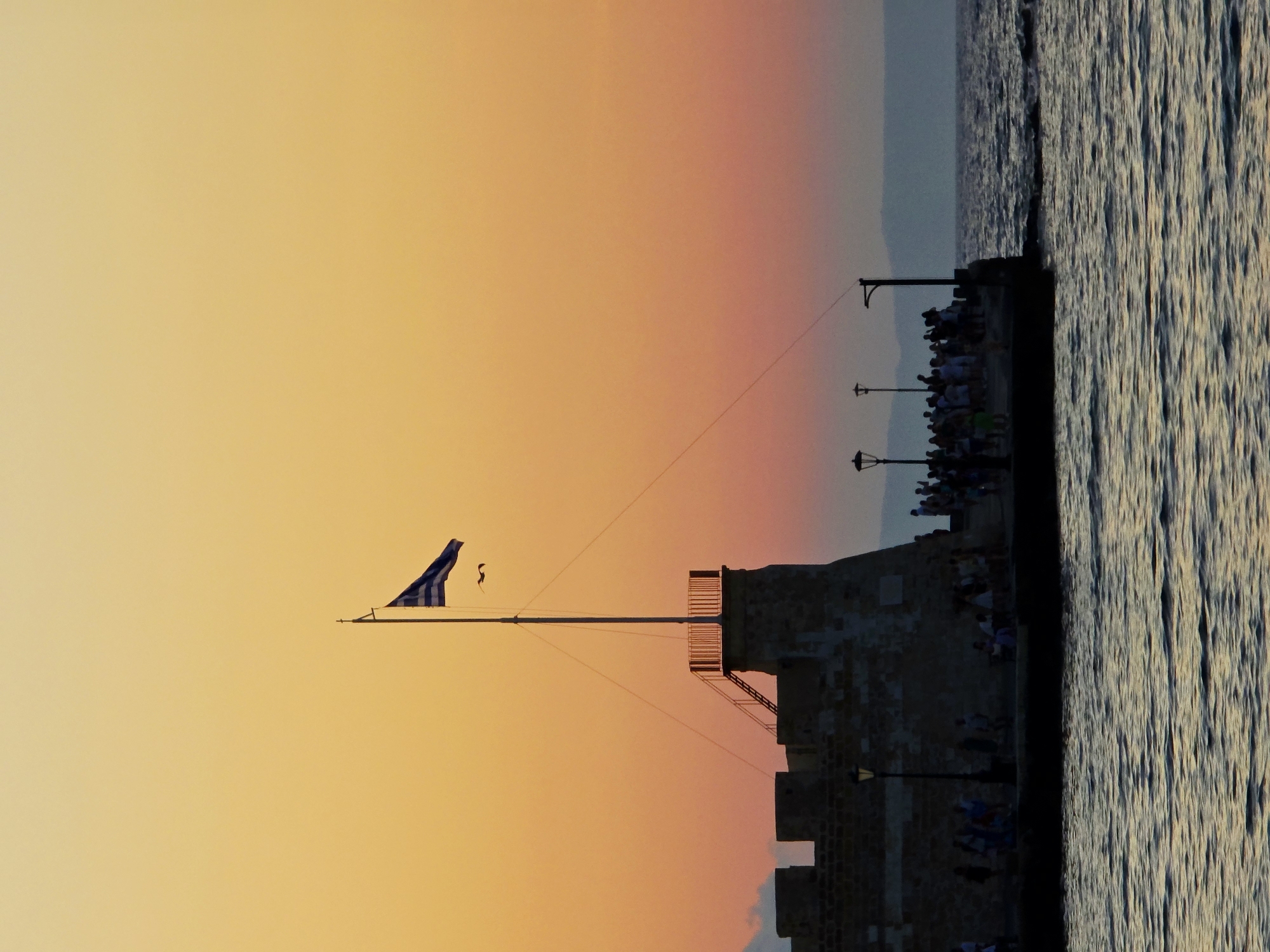 A wide shot captures a stone fortress, likely the Venetian harbor or Koules fortress, in Chania, Greece, at dusk. The sky is a gradient of warm orange and yellow, transitioning to a softer pink near the horizon, with distant mountains silhouetted. The sea in the foreground is dark and choppy, reflecting the sky's colors. A tall flagpole stands prominently on the fortress, flying a Greek flag. A flock of birds flies near the flag. Numerous people are silhouetted on the fortress walls and a pier extending into the water, some appearing to be observing the sunset or walking. Lampposts are visible along the pier. The stone structure of the fortress is detailed with crenellations and a small tower with stairs. The overall scene depicts a serene evening at a historical landmark.